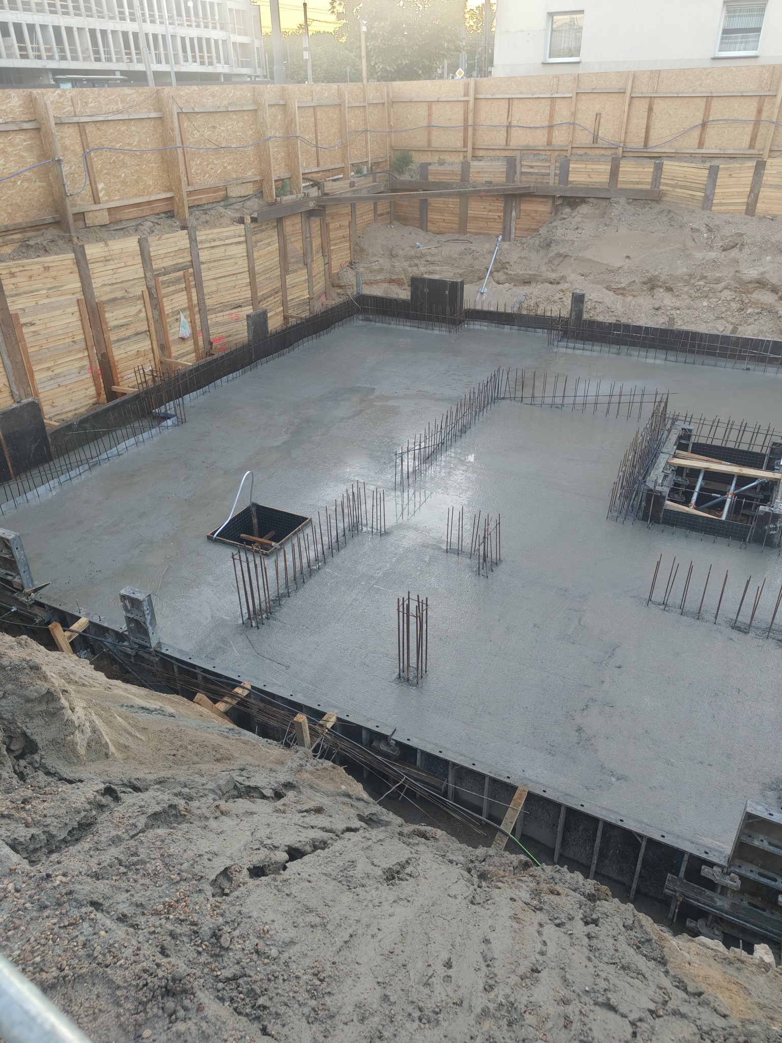 Zalany fundament z wystającymi prętami zbrojeniowymi i drewnianym szalunkiem. Widok z góry na wykopie zabezpieczonym drewnem i ziemią. W tle budynek.