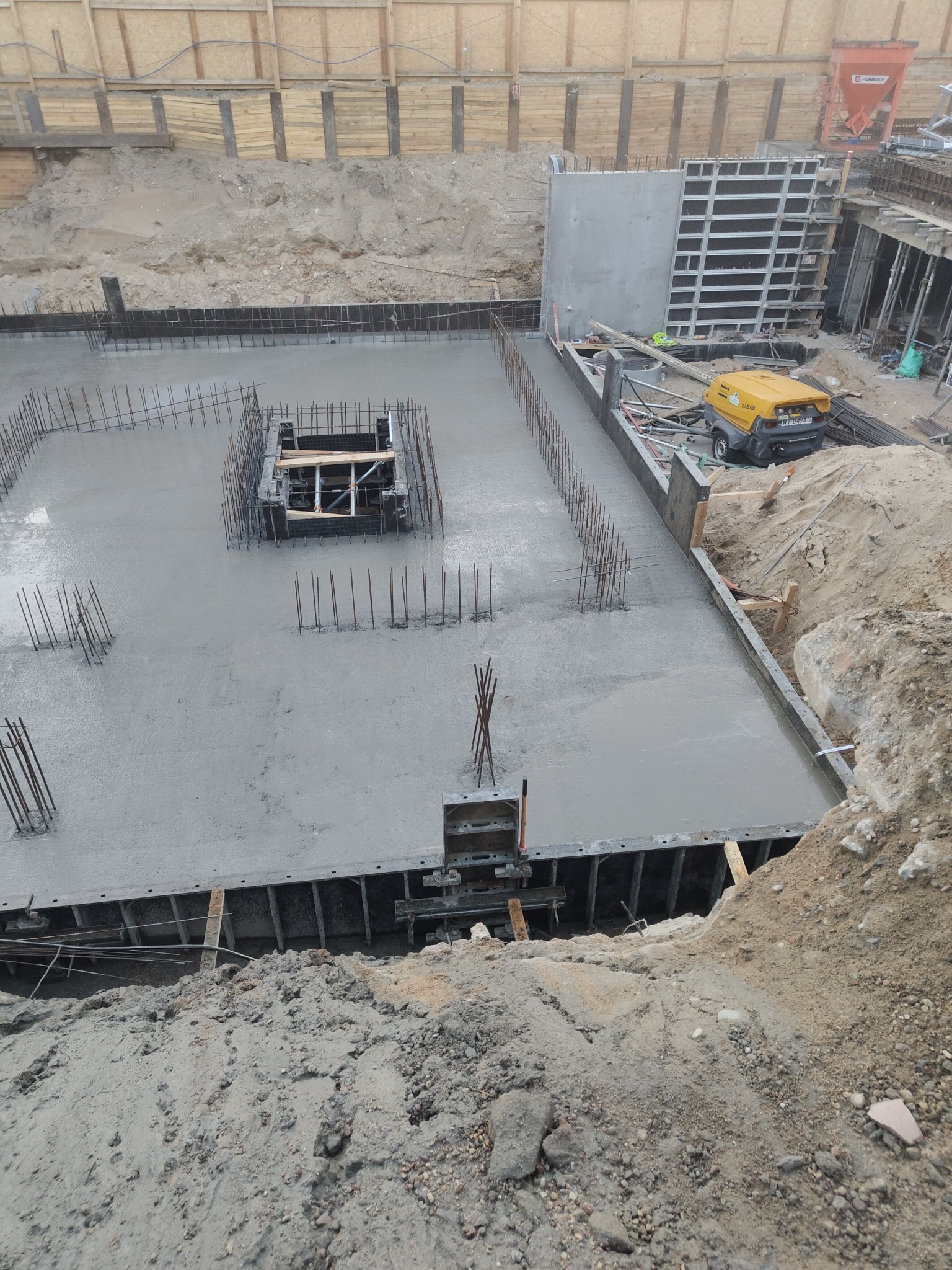 Zalany betonem fundament z wystającymi prętami zbrojeniowymi i szalunkami. Widoczny żółty agregat prądotwórczy i drewniane zabezpieczenia wykopu.
