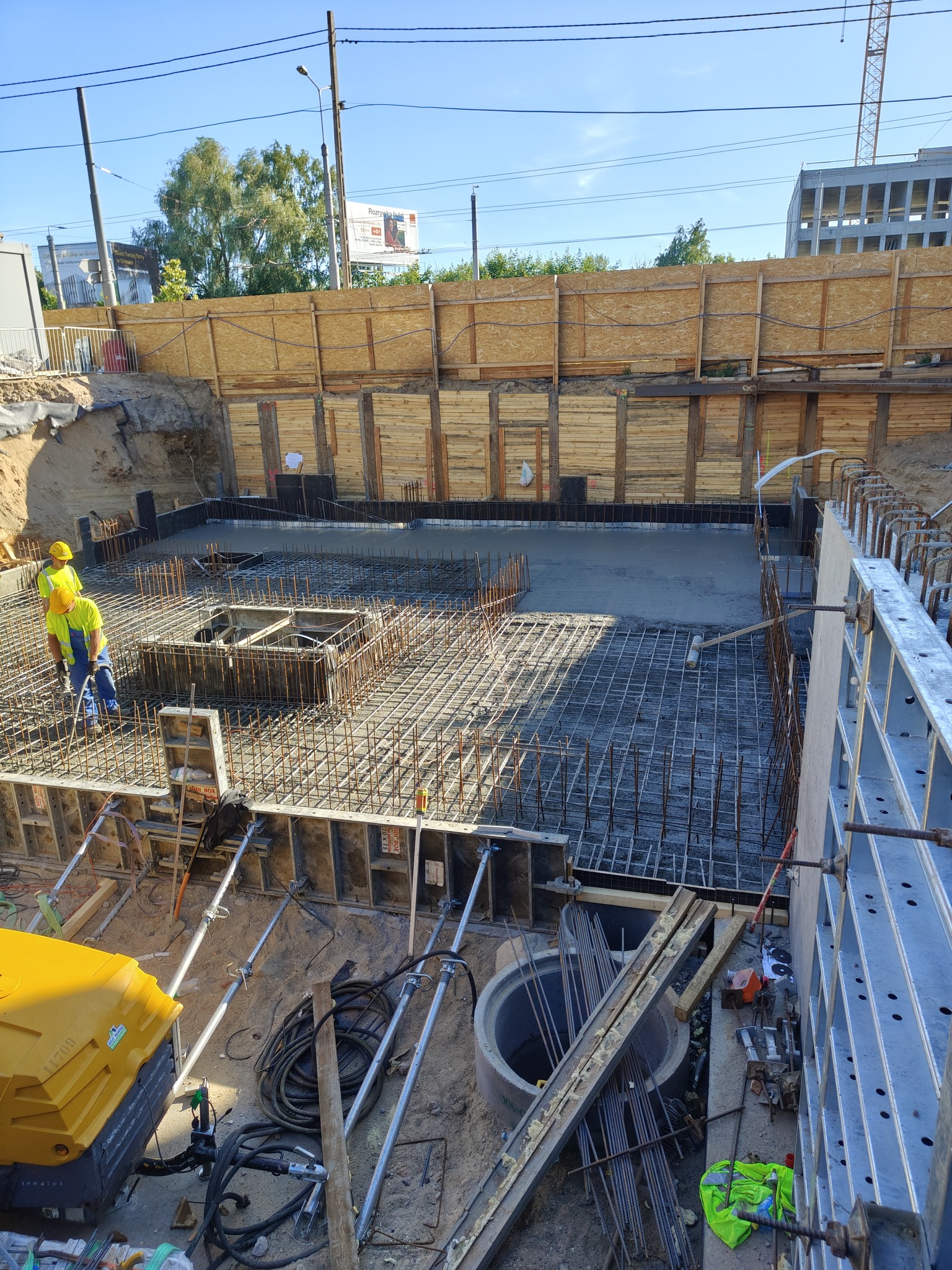 Zalewanie fundamentów na budowie. Widoczni pracownicy w żółtych kamizelkach, zbrojenie i szalunki. Ujęcie z góry na wykop z zabezpieczonymi ścianami.