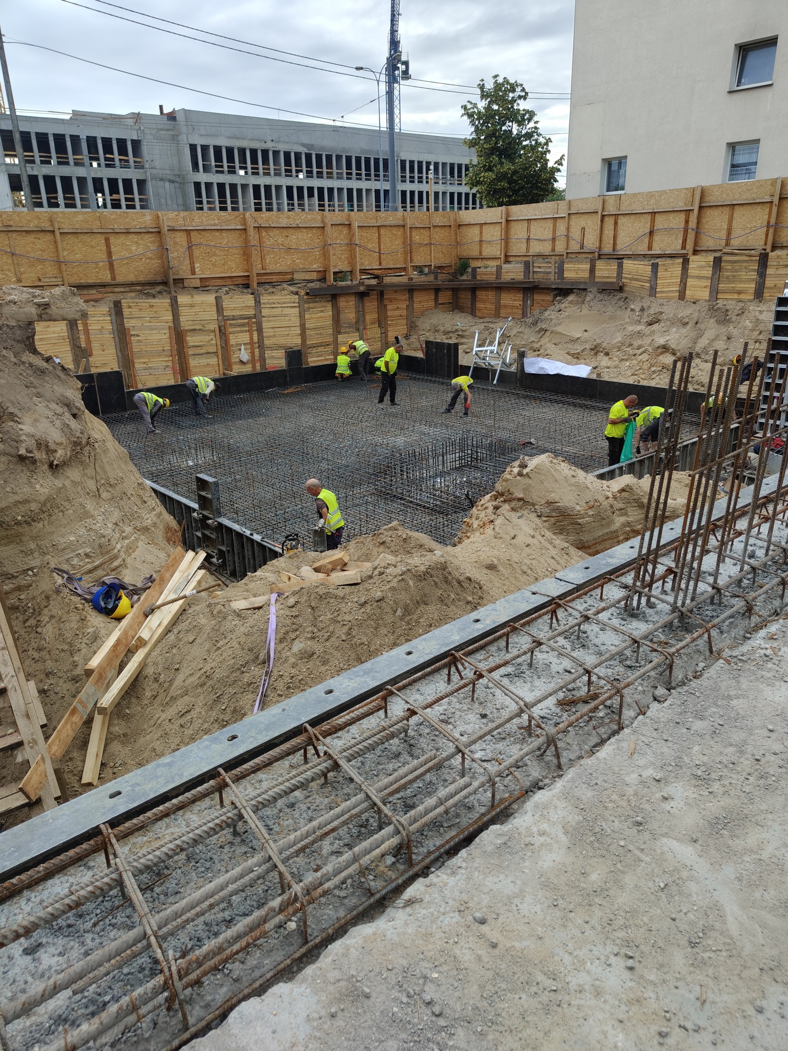 Prace zbrojarskie na budowie: robotnicy w żółtych kamizelkach układają stalowe pręty zbrojeniowe w wykopie fundamentowym, zabezpieczonym drewnianą szalunką.