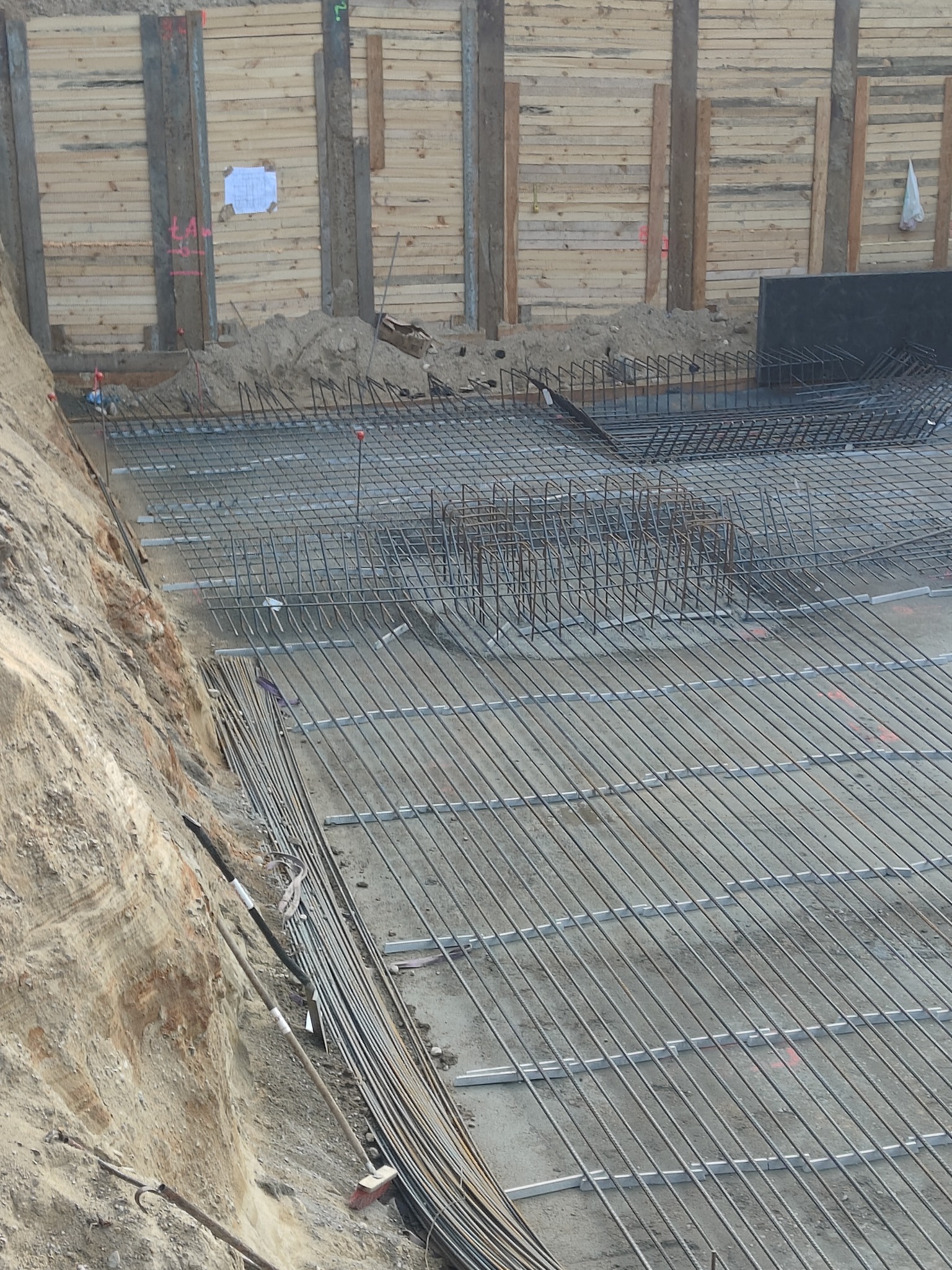 Zbrojenie fundamentów w wykopie zabezpieczonym drewnianą szalunką, widoczne pręty zbrojeniowe ułożone w siatkę i pionowe strzemiona, przygotowane do zalania betonem.