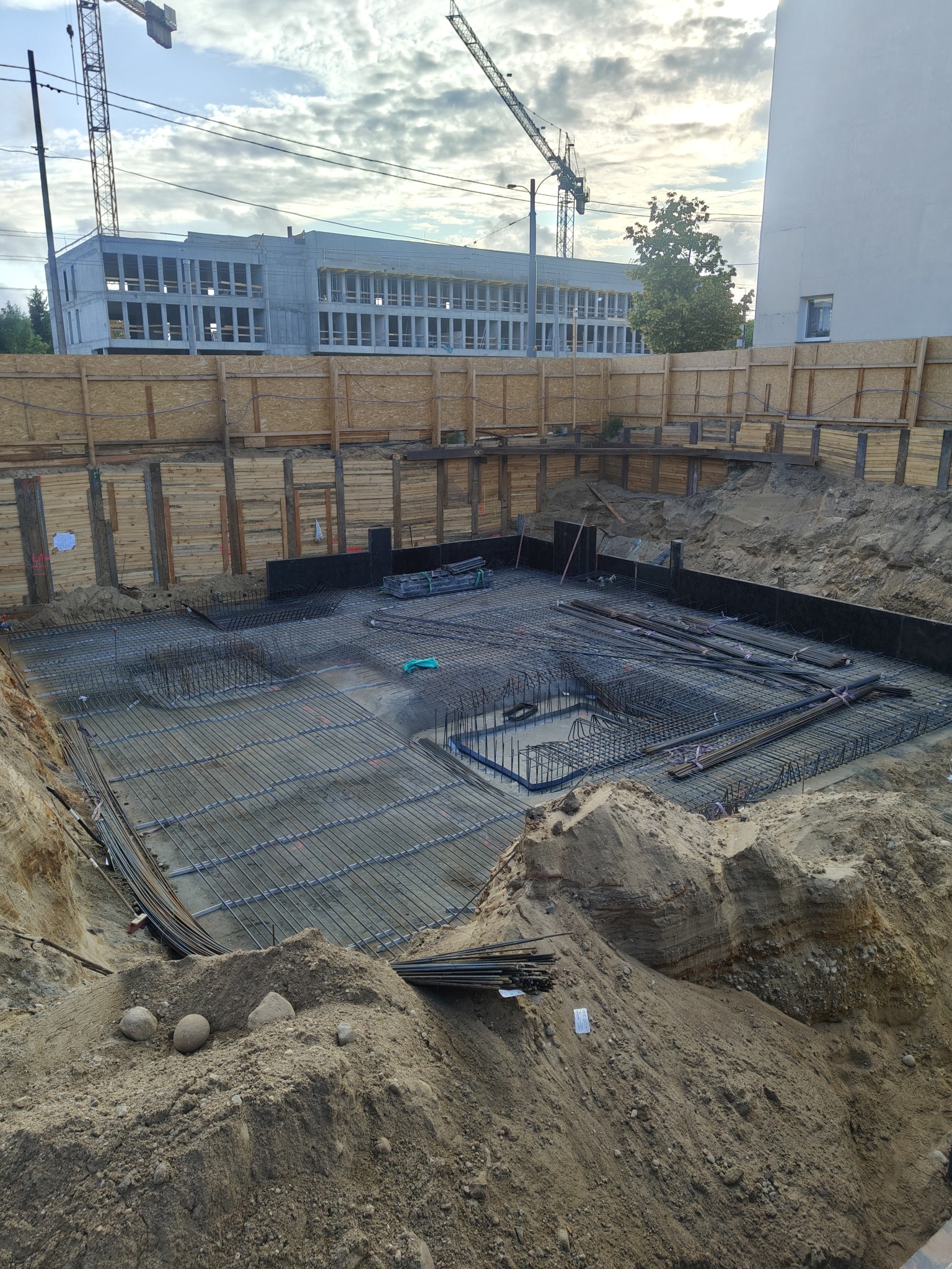 Zbrojenie fundamentów na budowie, widok z góry na wykop z przygotowanym zbrojeniem, w tle budynek w stanie surowym i żurawie budowlane.