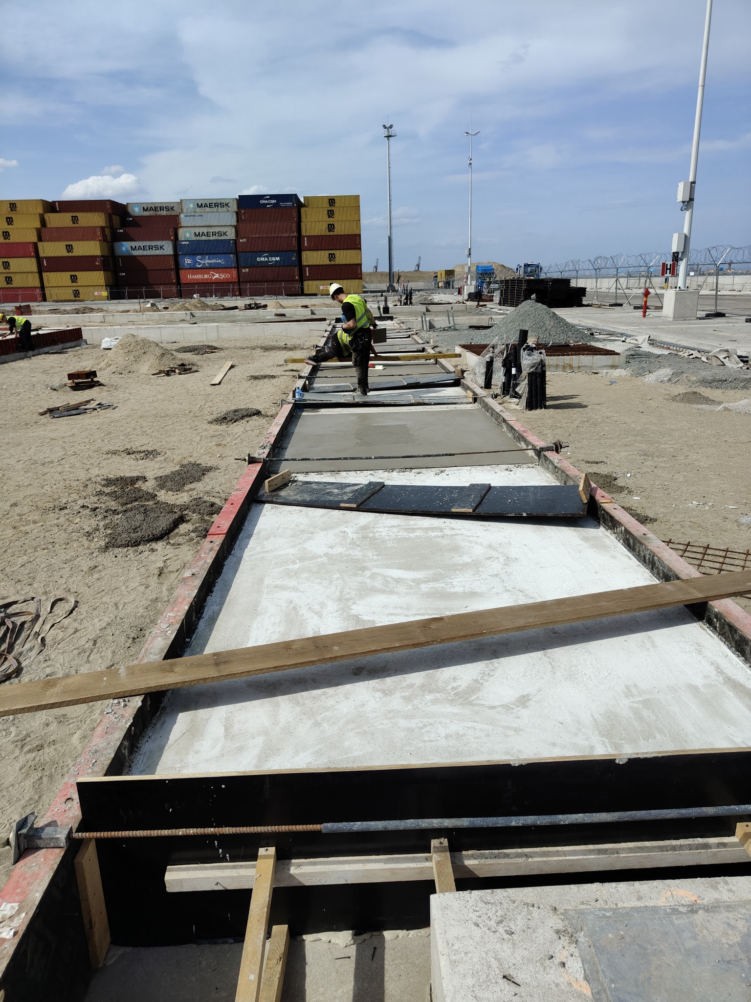 Wykonywanie betonowych fundamentów w szalunkach na placu budowy. W tle widoczne kontenery i pracownik w kamizelce odblaskowej. Ujęcie z góry.