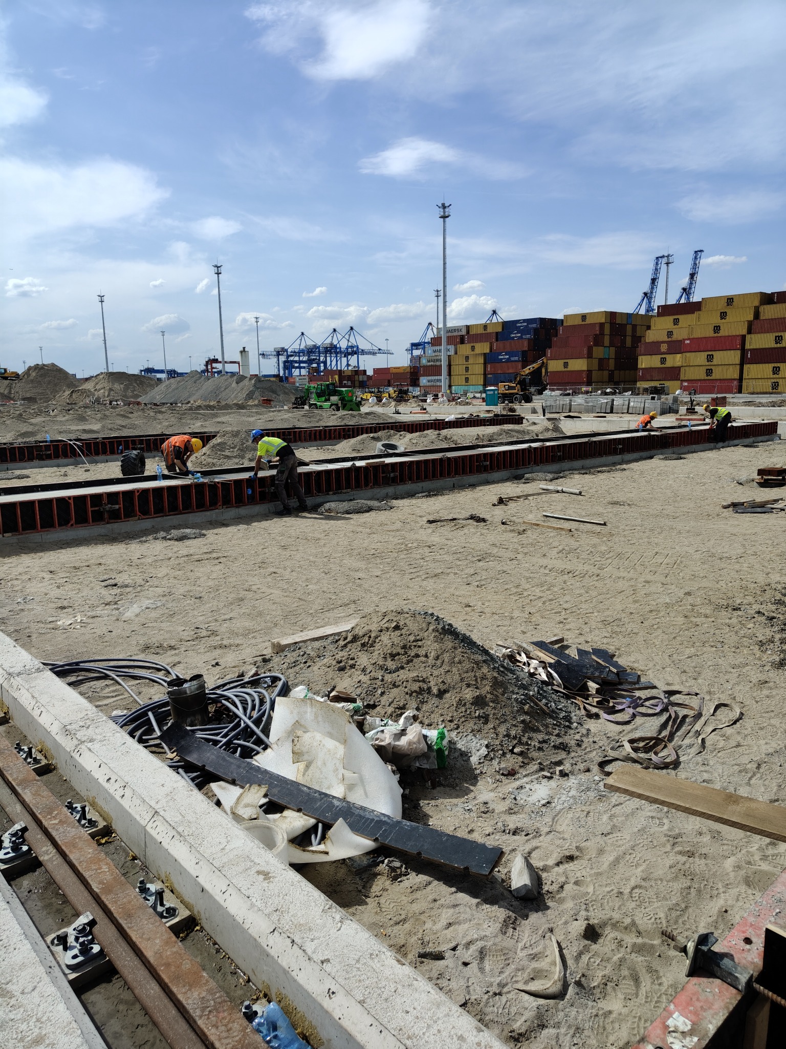 Prace zbrojarskie na budowie: pracownicy w kaskach montują szalunki pod fundamenty, w tle widoczne dźwigi portowe i kontenery. Słoneczny dzień, plac budowy.