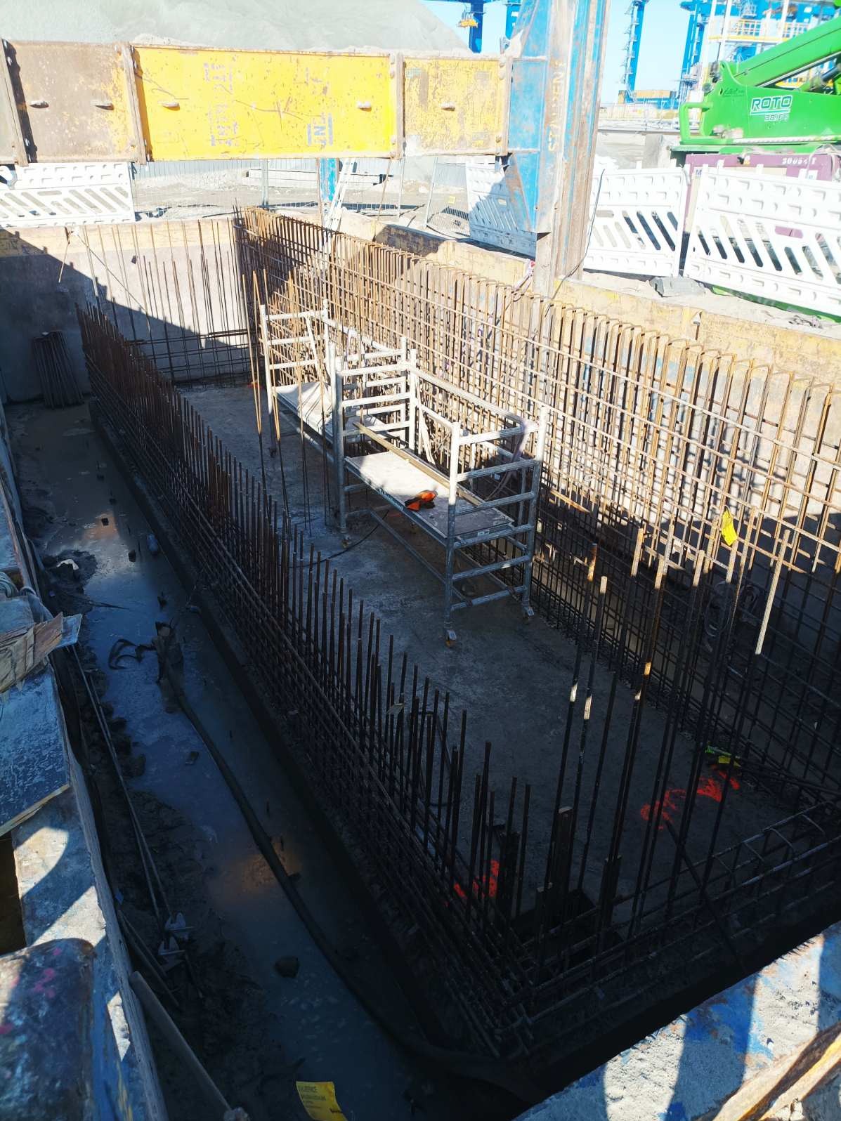 Zbrojenie fundamentów w wykopie, widok z góry. Stalowe pręty tworzą konstrukcję szkieletową. W tle widoczny sprzęt budowlany i elementy konstrukcyjne.