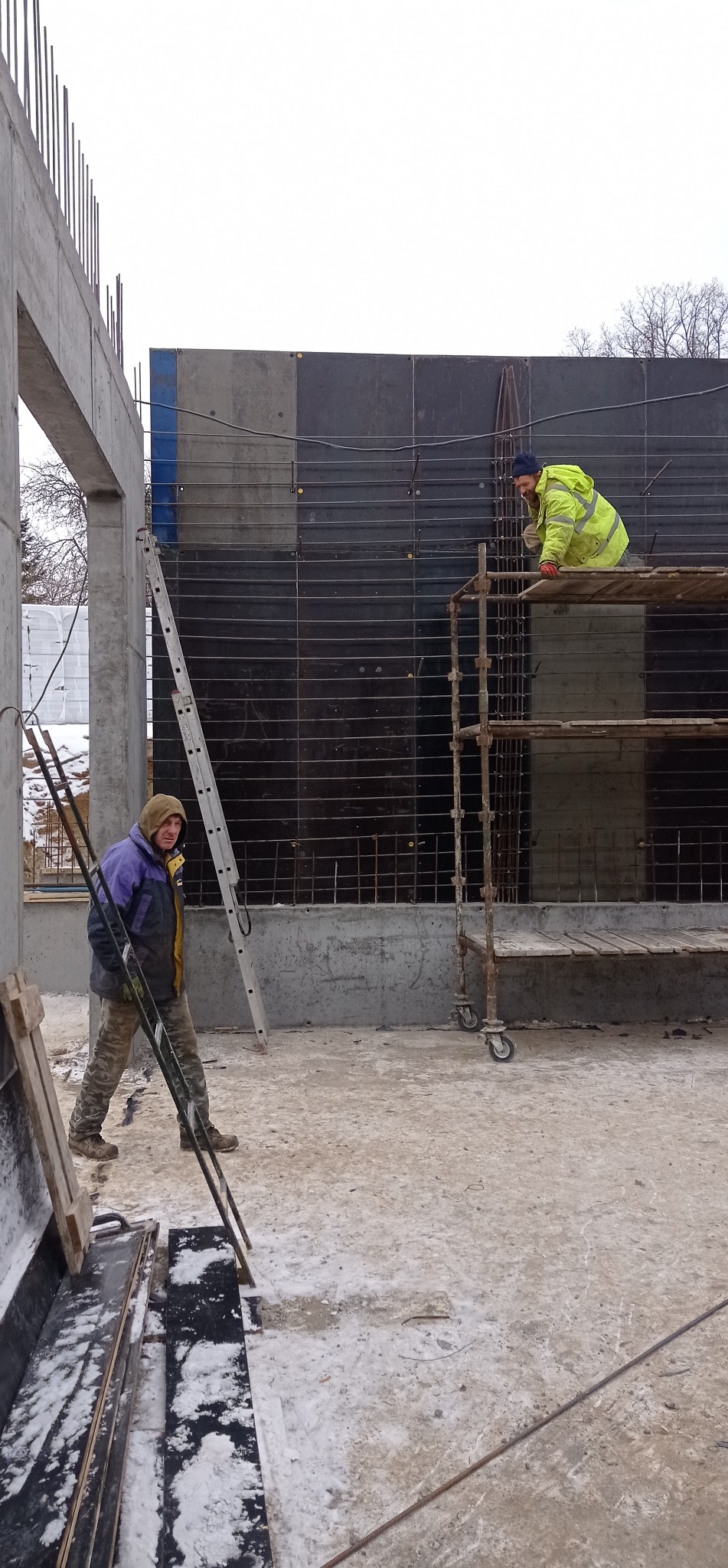 Dwóch pracowników w odzieży roboczej na placu budowy, jeden na rusztowaniu przy szalunku, drugi przenosi drabinę. Widoczne zbrojenia i betonowe elementy konstrukcyjne.