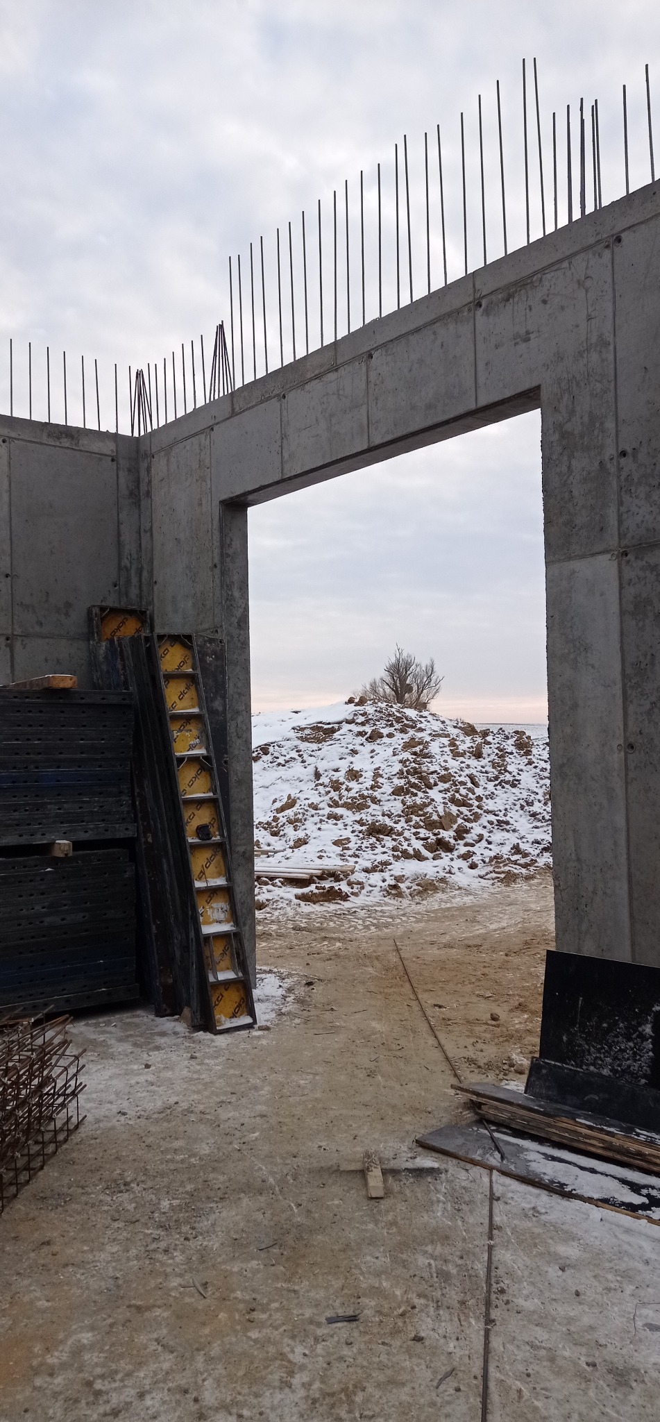 Surowa konstrukcja żelbetowa z wystającymi prętami zbrojeniowymi, widok przez otwór na zasypaną śniegiem ziemię. Szalunki czekają na użycie.