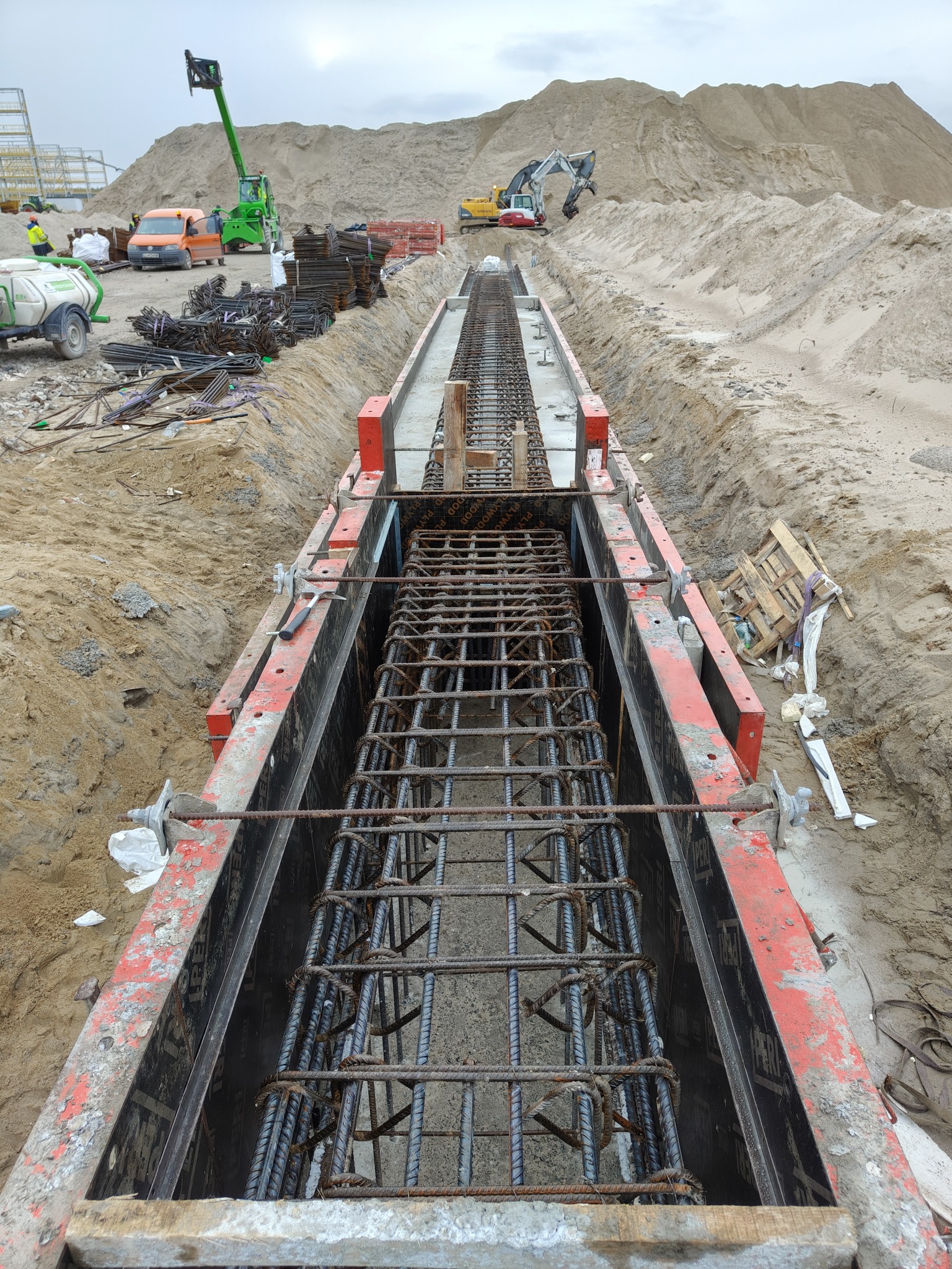 Zbrojenie w szalunku, widok z góry na przygotowany wykop. W tle hałda piasku i sprzęt budowlany. Drobne elementy zbrojeniowe poza główną konstrukcją.