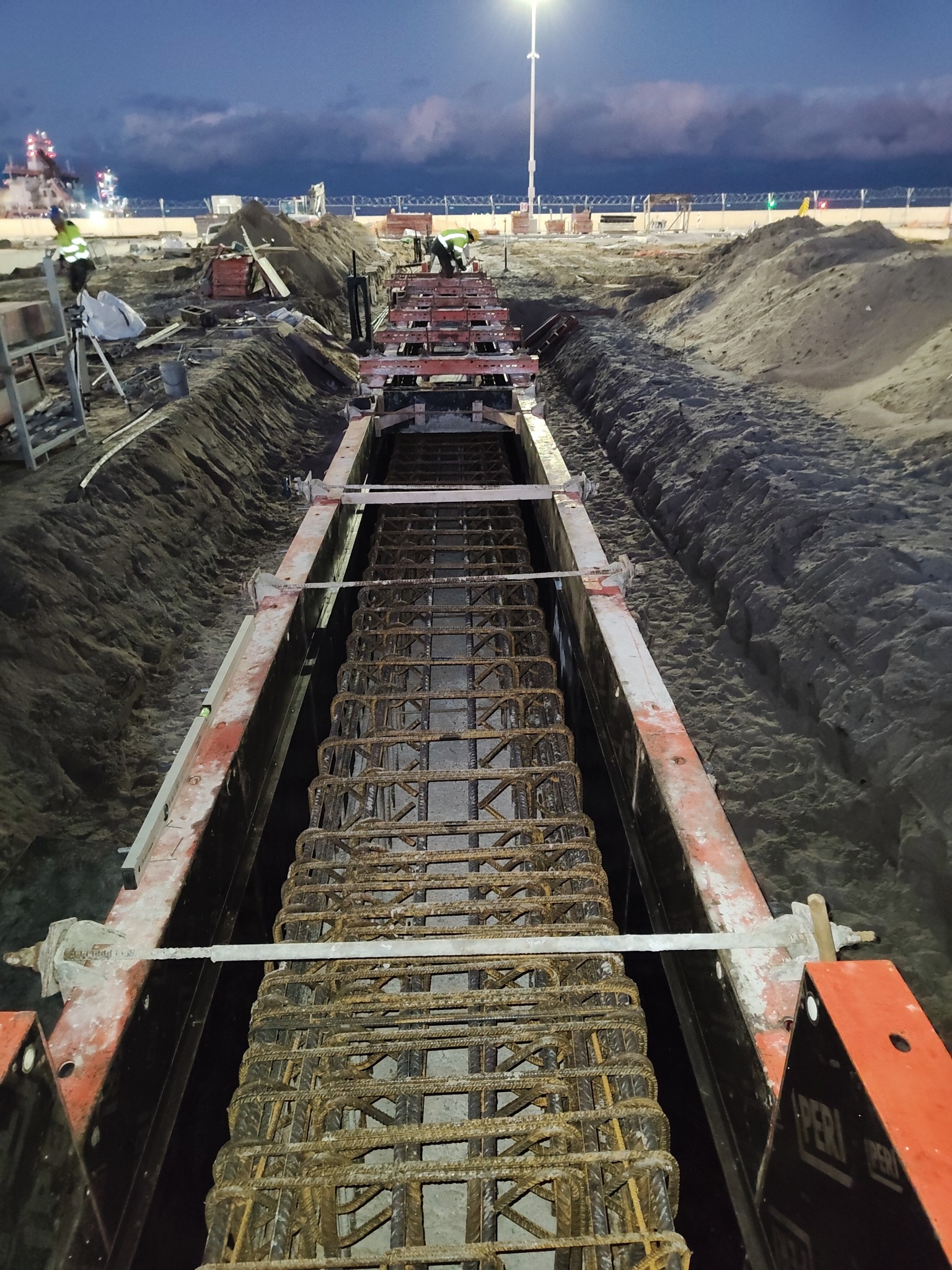 Zbrojenie fundamentów w wykopie, widok z góry. Metalowa konstrukcja zbrojeniowa w szalunku, przygotowana do zalania betonem. Prace ziemne w tle, oświetlenie placu budowy.