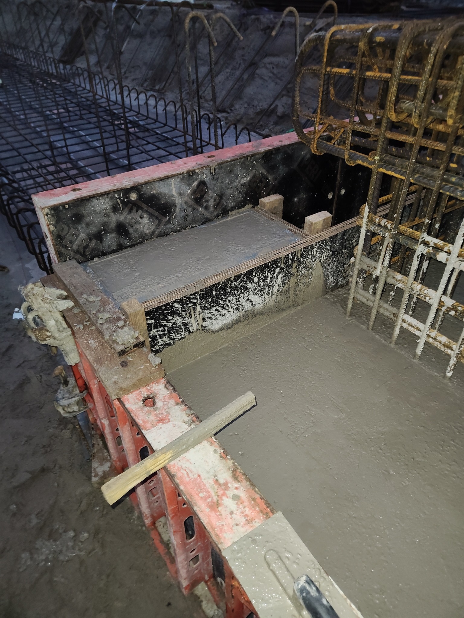 Zalewanie betonem zbrojonej konstrukcji szalunkowej. Widoczne detale zbrojenia i świeżo wylany beton w szalunku z elementami drewnianymi i metalowymi.