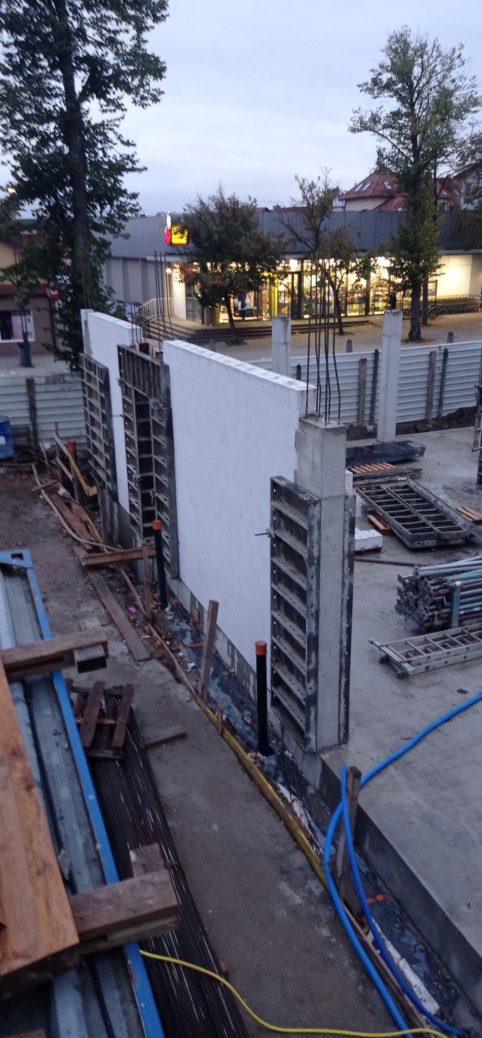 Budowa ściany z bloczków betonowych z szalunkiem i wystającymi prętami zbrojeniowymi. Widoczne elementy konstrukcyjne i narzędzia budowlane na placu budowy.