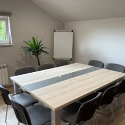 Sala konferencyjna z dużym stołem, krzesłami, flipchartem i rośliną doniczkową przy oknie. Jasne wnętrze, parkiet na podłodze, grzejnik pod oknem.