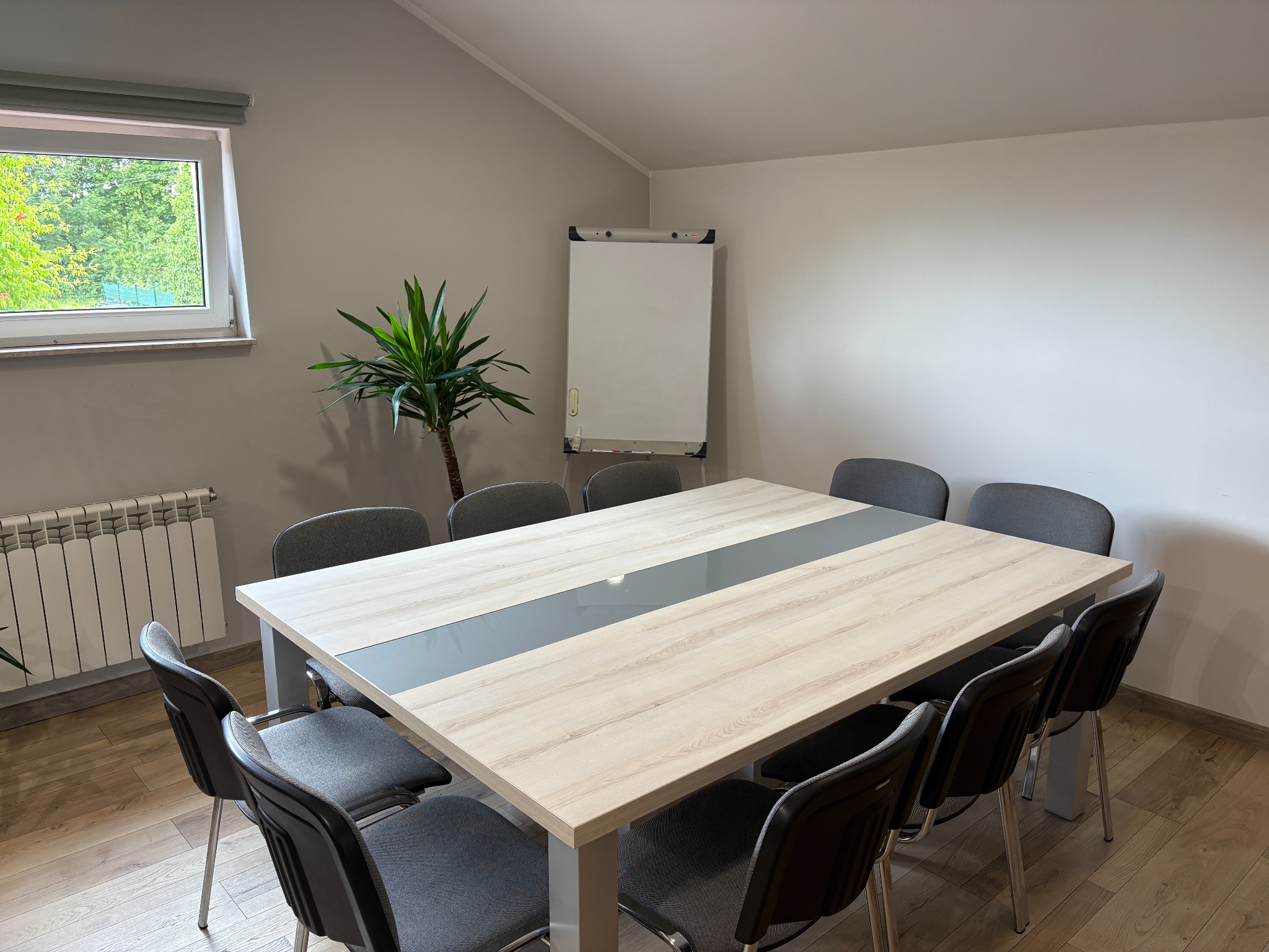 Sala konferencyjna z dużym stołem, krzesłami, flipchartem i rośliną doniczkową przy oknie. Jasne wnętrze, parkiet na podłodze, grzejnik pod oknem.