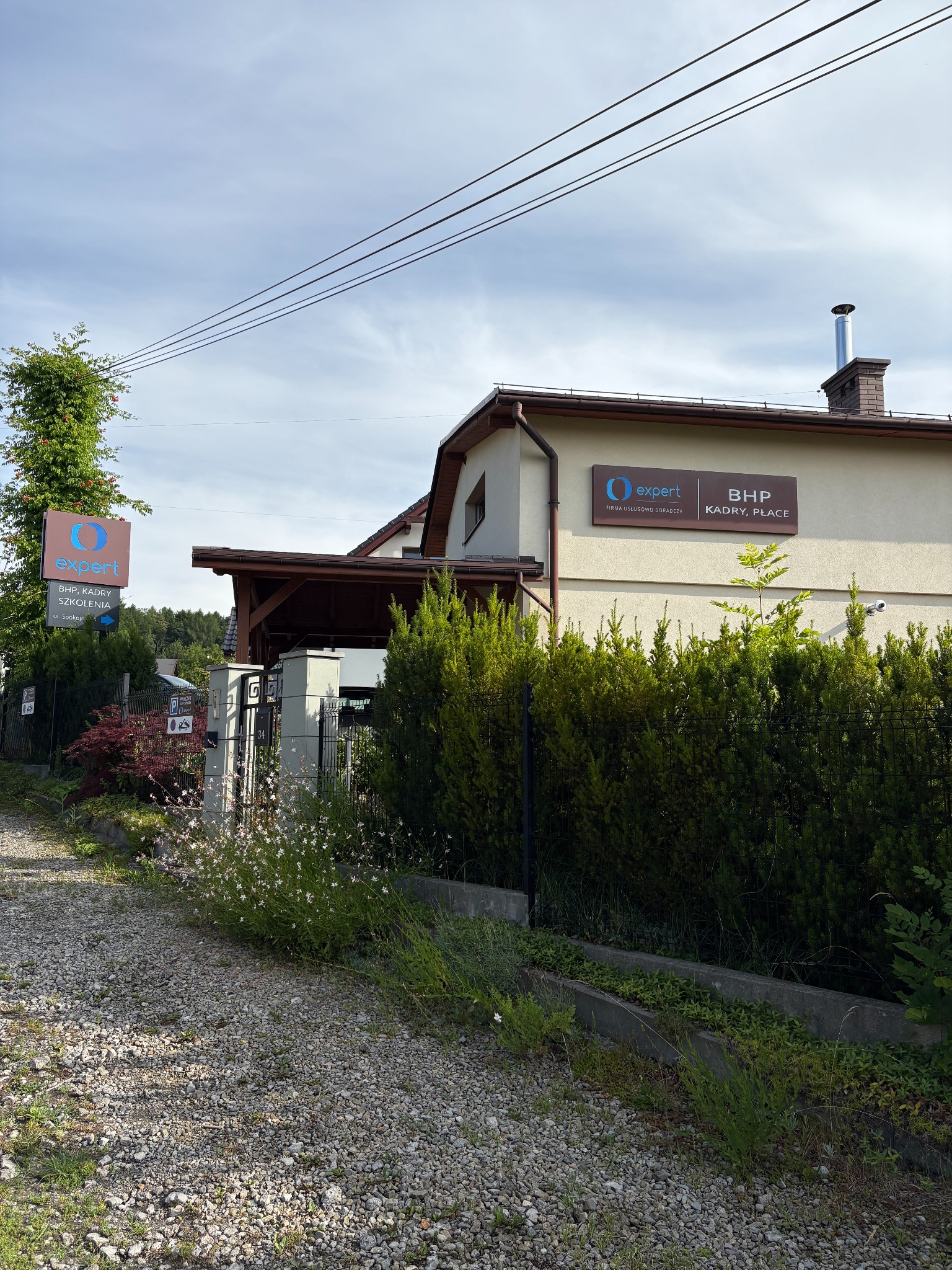 Budynek firmy doradczej Expert z tablicami informacyjnymi BHP, Kadry, Szkolenia. Widok z drogi szutrowej, zieleń wokół, pochmurne niebo.