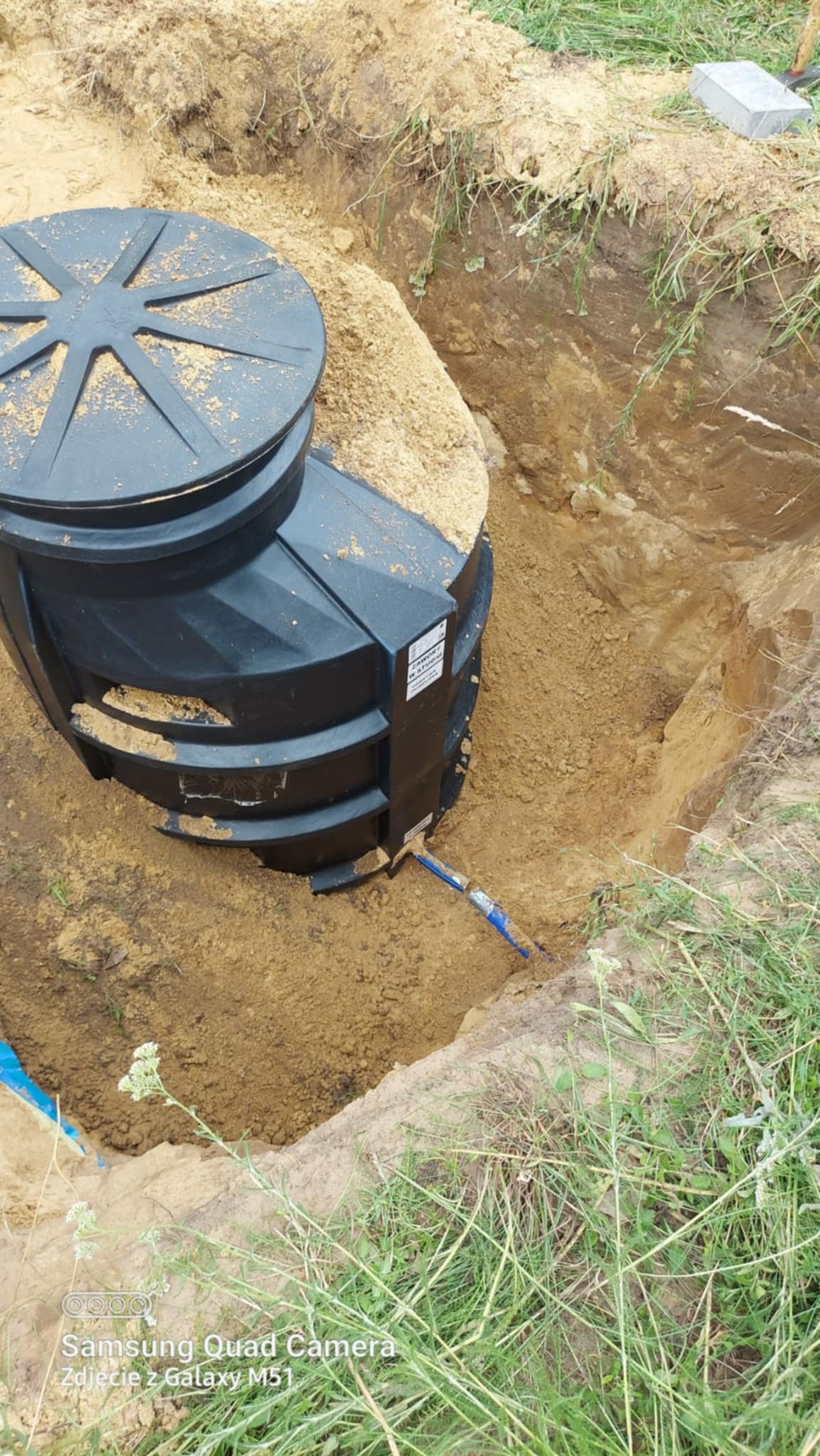 Czarny, plastikowy szambon betonowy częściowo zasypany piaskiem w wykopie, widoczny fragment niebieskiej rury. Ujęcie z góry, trawa w tle.