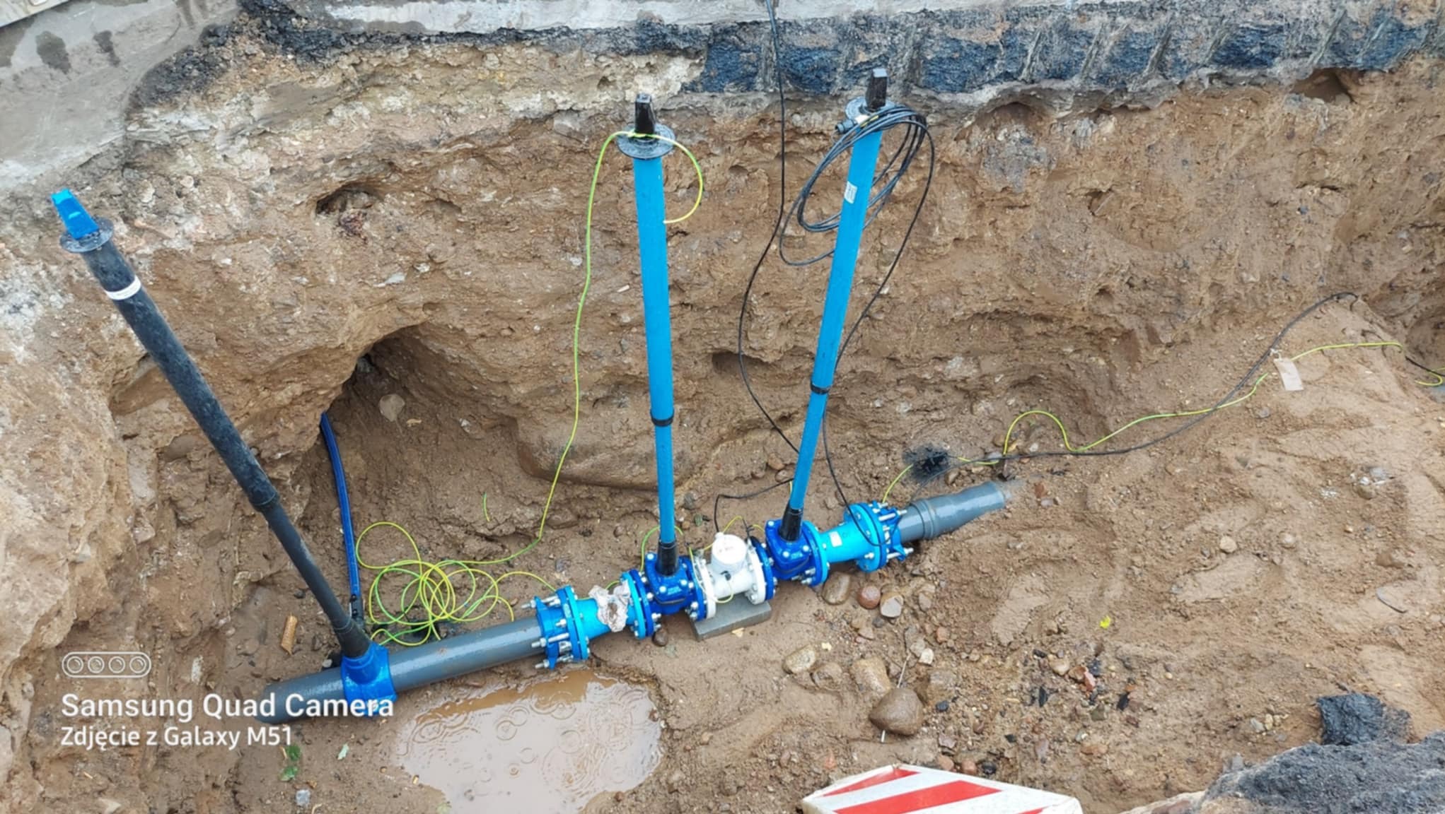 Wykop z widocznymi rurami kanalizacyjnymi z niebieskimi elementami łączeniowymi, wystającymi pionowo rurami i kablami. W tle ściana wykopu z ziemi.