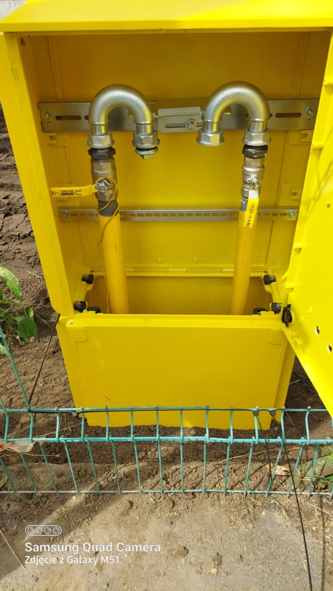 Żółta skrzynka gazowa z zaworami i rurami, widoczne elementy instalacji gazowej na tle ziemi i zielonego ogrodzenia.