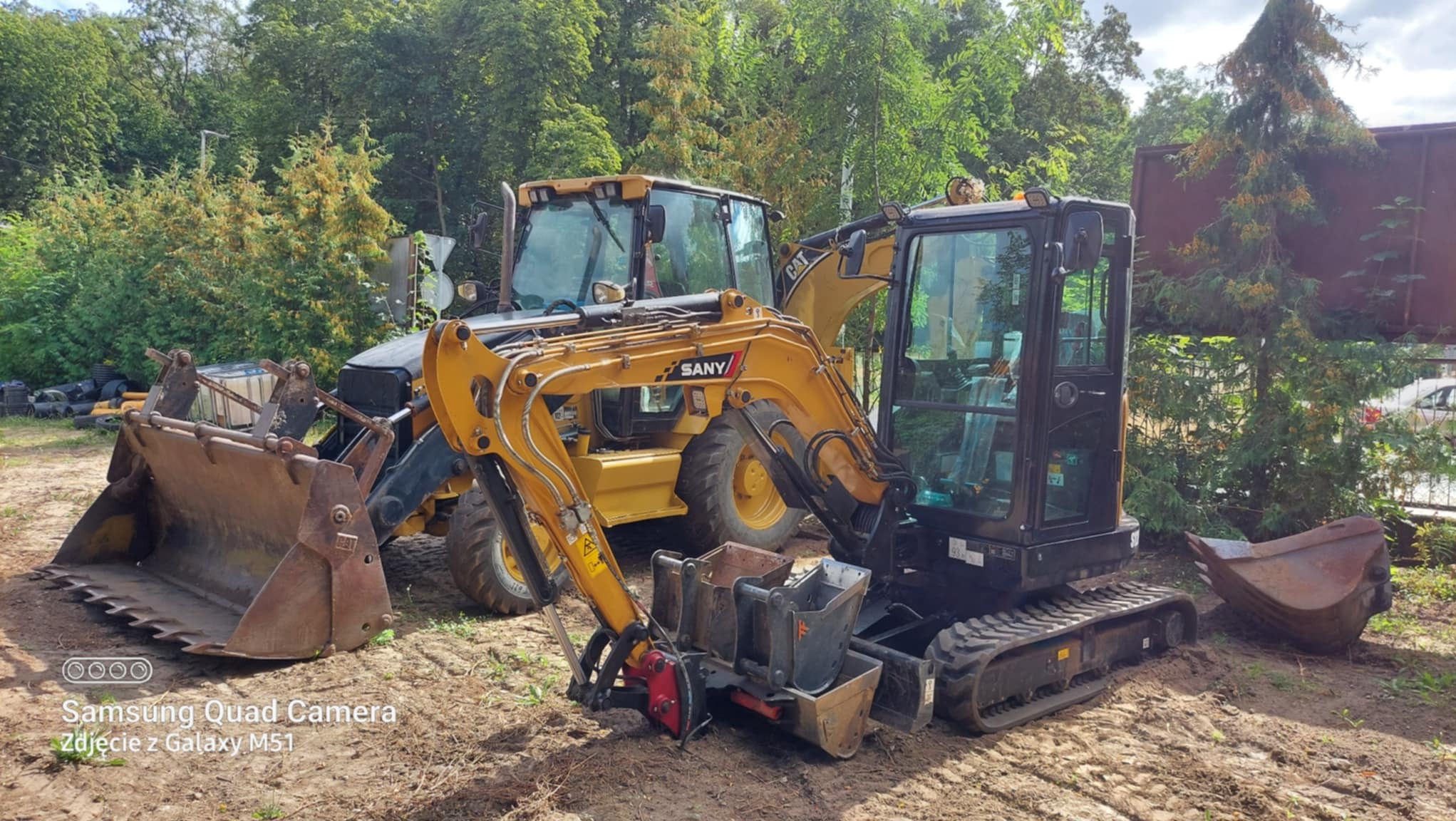 Dwie maszyny budowlane: żółta ładowarka CAT i czarna mini-koparka SANY stoją na ziemi, otoczone zielenią. Widoczne detale maszyn i narzędzia.
