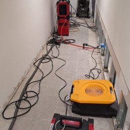 SKM-BUD Łukasz Kowalczyk - Sprzęt do osuszania w trakcie pracy w pomieszczeniu. Widoczne czerwone i żółte urządzenia, kable i metalowe profile ścian. Szare płytki podłogowe.