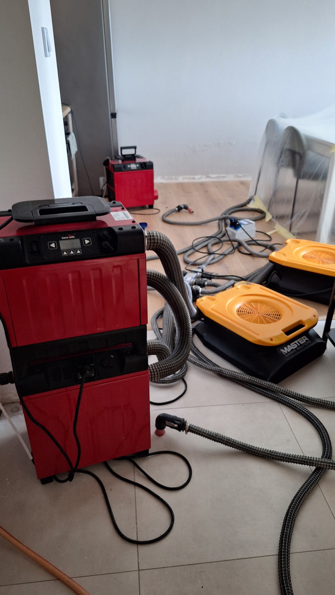 Sprzęt do osuszania: czerwony osuszacz powietrza Corroventa i dwa żółte osuszacze Master Climate Solutions z szarymi wężami na podłodze z płytek w trakcie pracy.