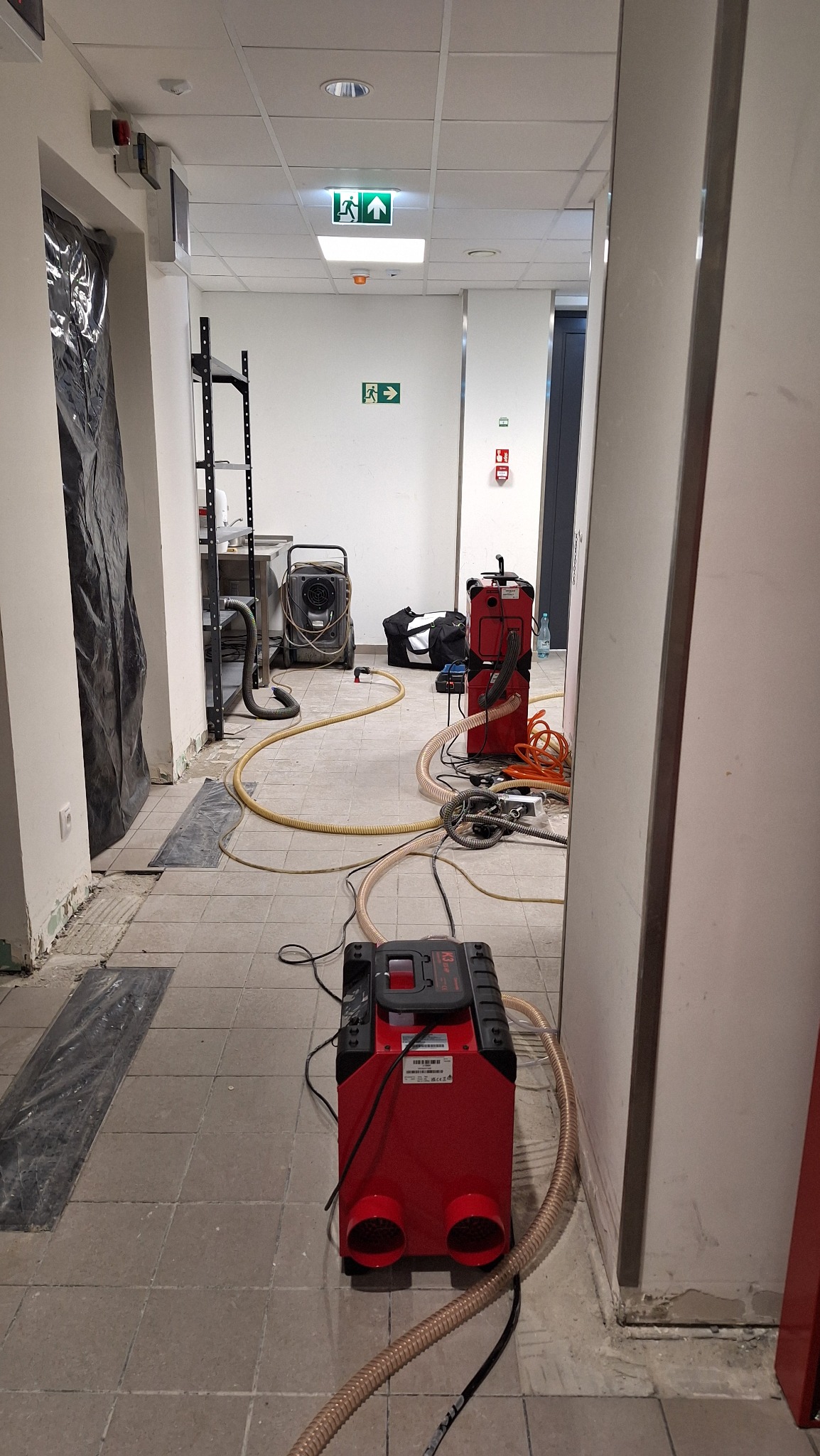Korytarz w trakcie prac. Widoczne urządzenia do osuszania podłogi i ścian, węże oraz zabezpieczone folią fragmenty posadzki. Oznakowanie ewakuacyjne w tle.