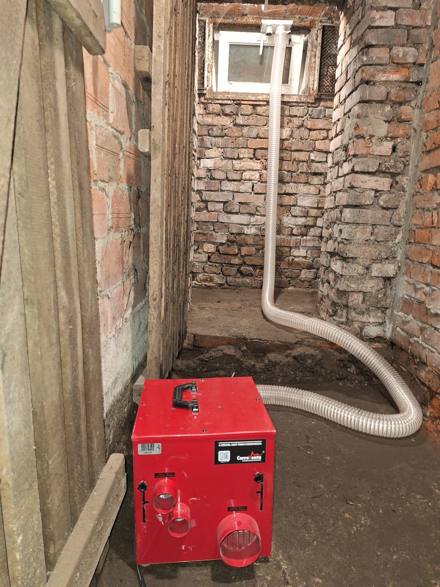 Czerwony osuszacz kondensacyjny Corroventa z podłączonym wężem w pomieszczeniu z ceglanymi ścianami, osuszanie murów.