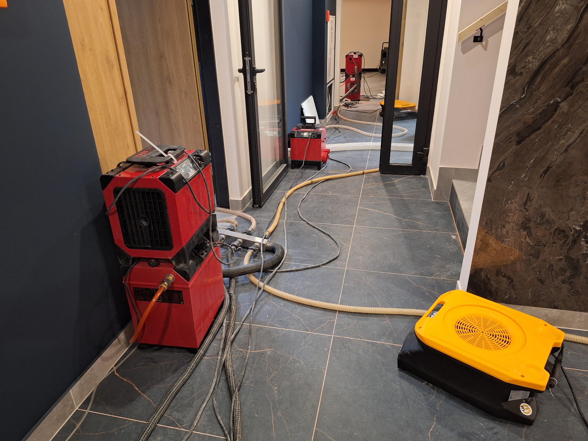 Czerwony sprzęt do osuszania stoi na ciemnej, marmurkowej podłodze w korytarzu. W tle widoczne kolejne urządzenia i otwarte drzwi.