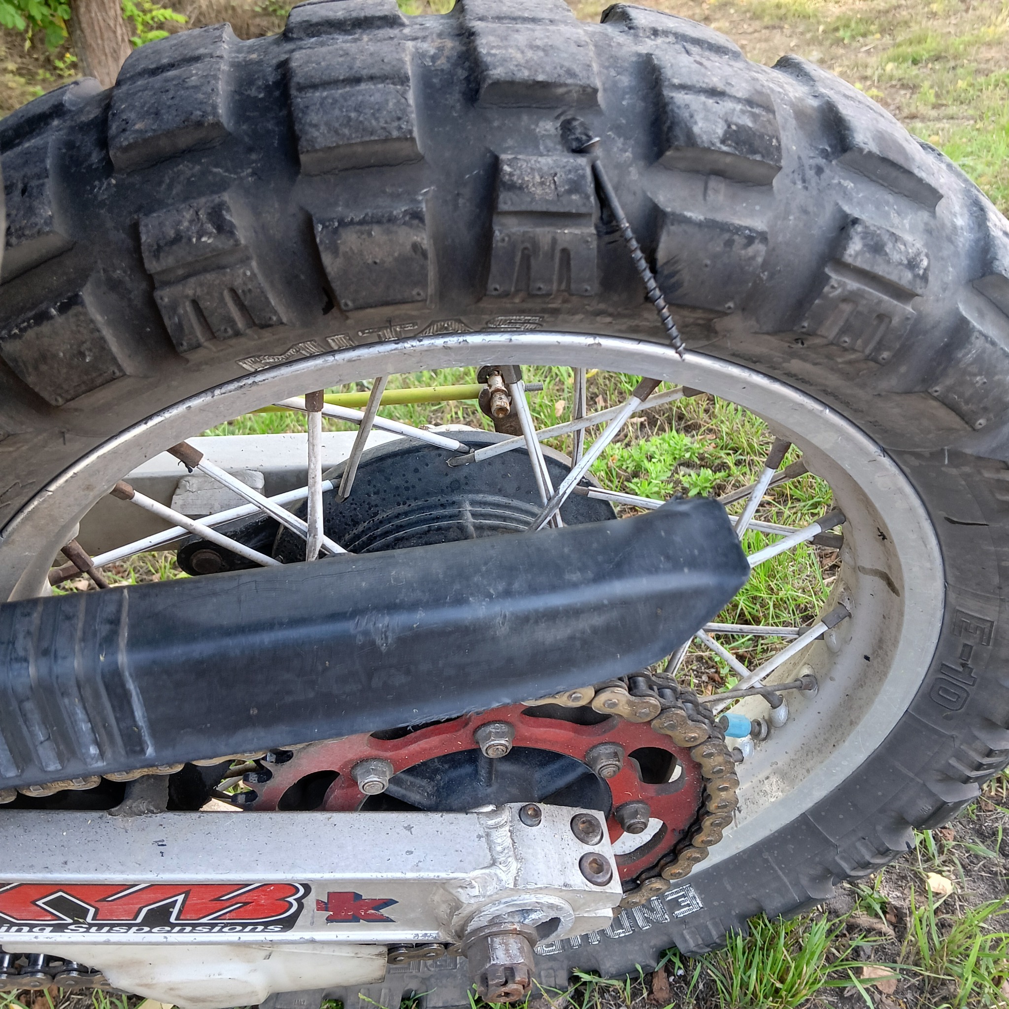 Bieżnikowana opona motocyklowa z widocznym gwoździem, szprychy koła i łańcuch napędowy. Widok z bliska na tle trawy i drzewa.