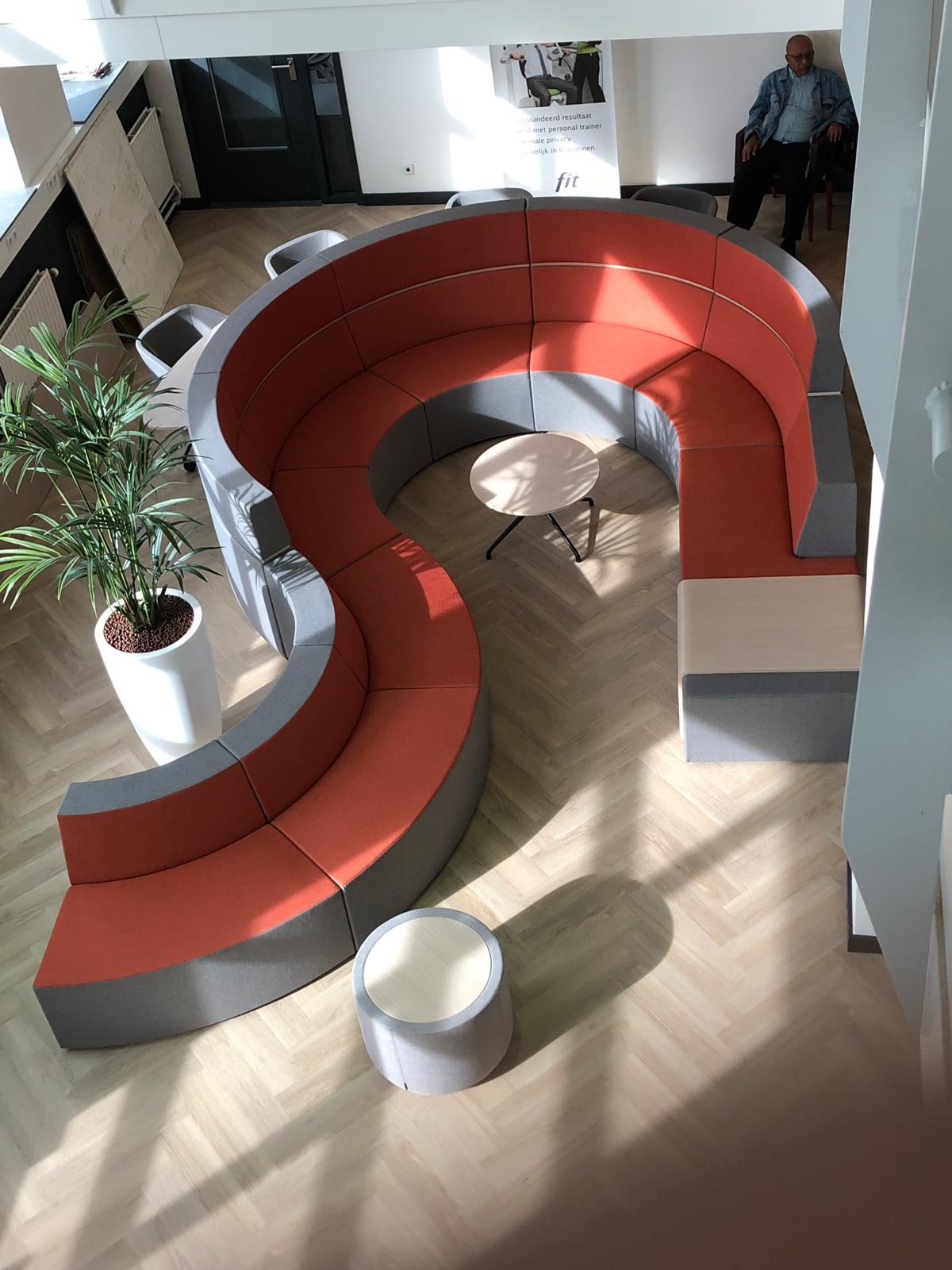 Widok z góry na modułową sofę w kształcie litery U, z pomarańczowymi siedziskami i szarą podstawą, ustawioną w jasnym pomieszczeniu z drewnianą podłogą w jodełkę.