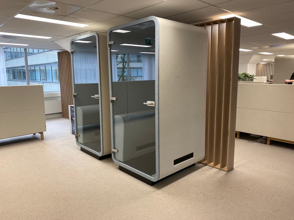Dwie nowoczesne, tapicerowane budki akustyczne z przeszklonymi drzwiami w jasnym biurze. Widoczne elementy tapicerki i metalowe uchwyty. Minimalistyczny design.