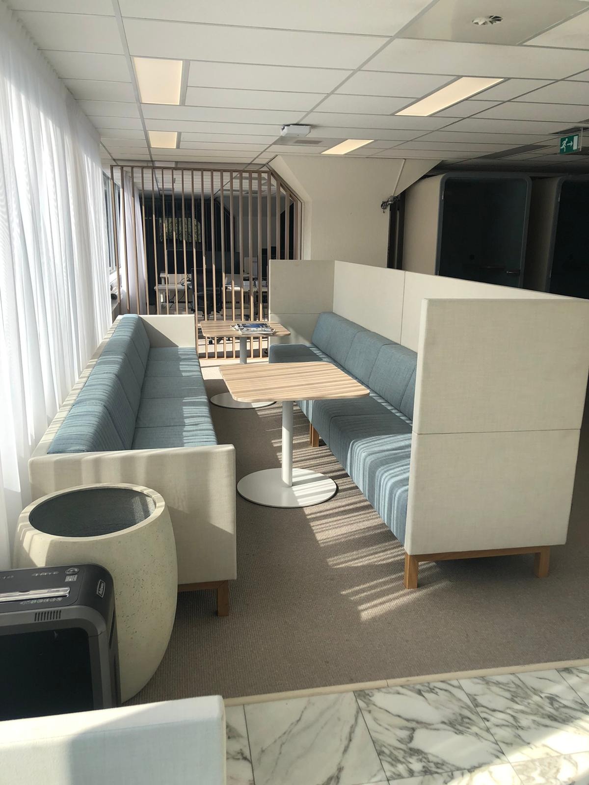 Nowoczesna strefa relaksu z tapicerowanymi sofami w odcieniach błękitu i beżu, oddzielona panelami, z drewnianymi stolikami na białych, okrągłych podstawach.