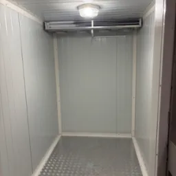 Wnętrze chłodni z metalową podłogą i panelem chłodniczym pod sufitem, oświetlone lampą. Widok z perspektywy wejścia, białe ściany z blachy trapezowej.