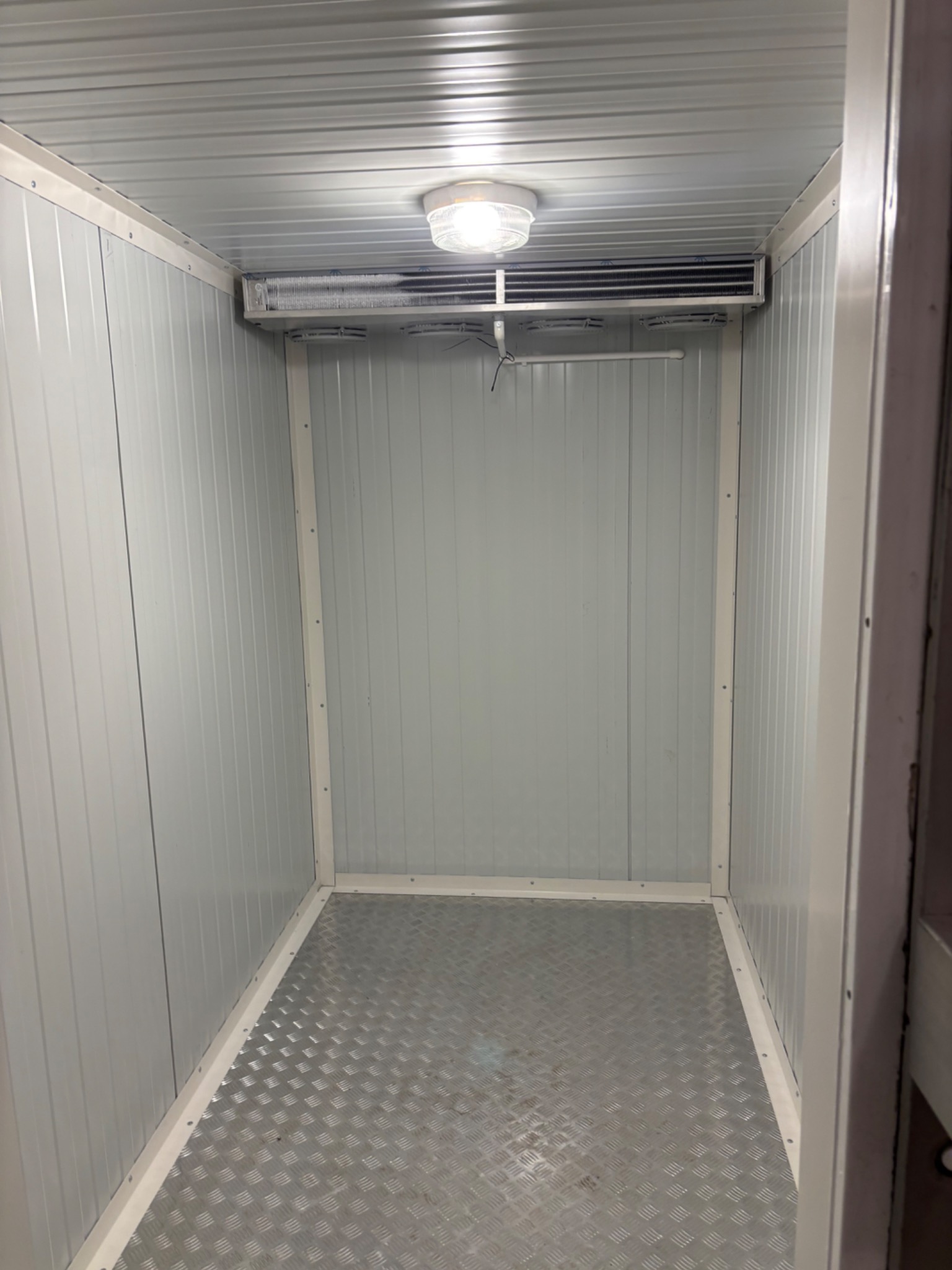 Wnętrze chłodni z metalową podłogą i panelem chłodniczym pod sufitem, oświetlone lampą. Widok z perspektywy wejścia, białe ściany z blachy trapezowej.