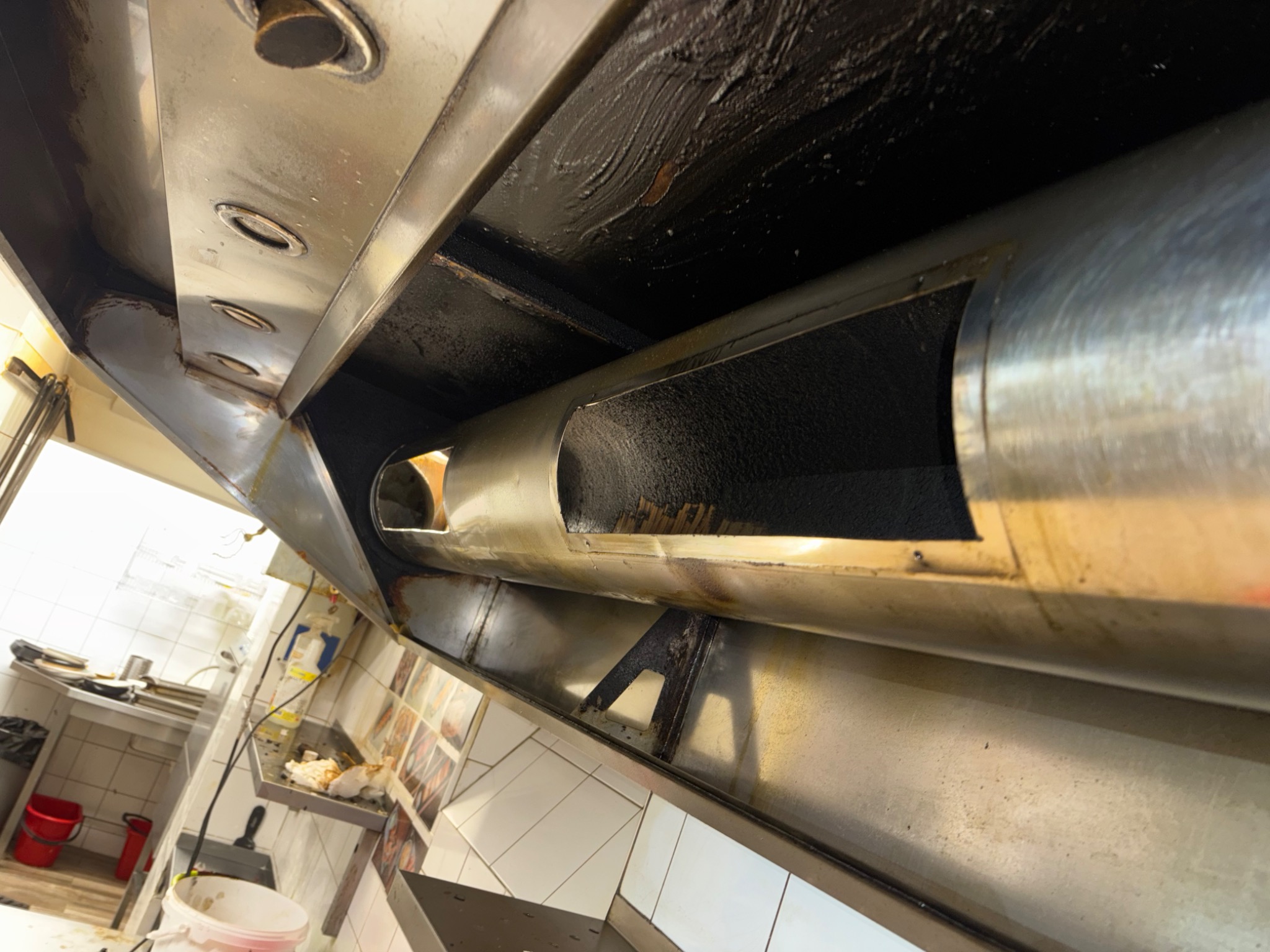 Zabrudzony okap kuchenny ze stali nierdzewnej w restauracji, widok od dołu, ukazujący tłuste osady i konstrukcję. Widoczne płytki i wyposażenie kuchenne w tle.