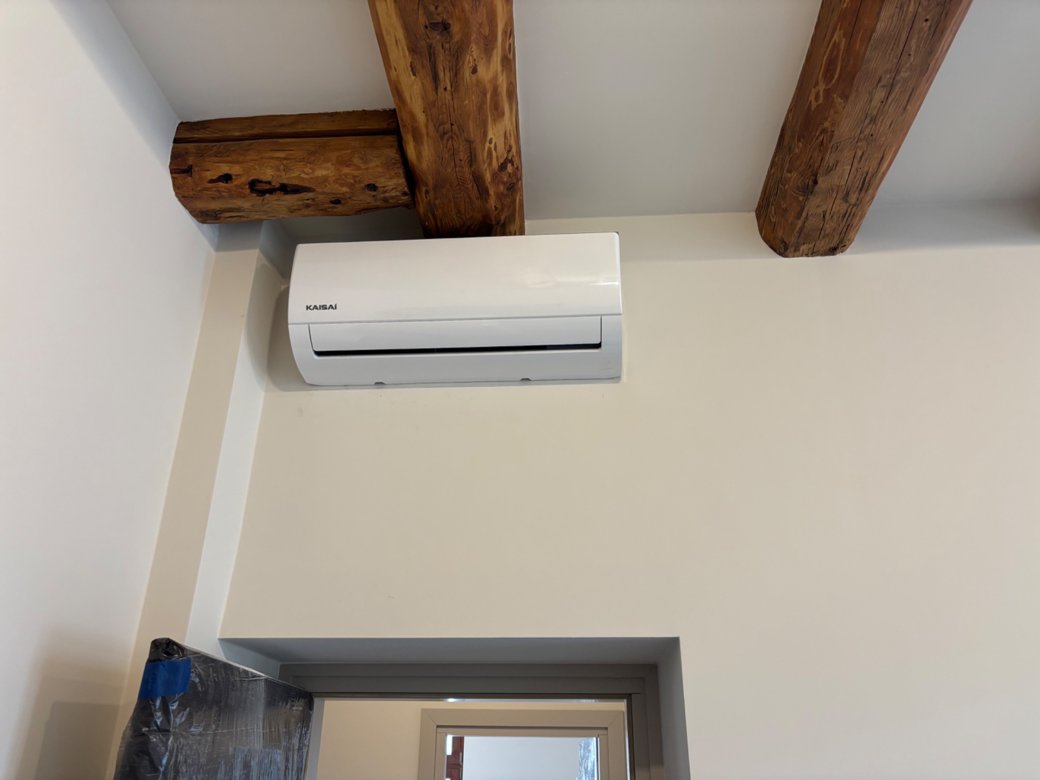 Biała jednostka klimatyzacji Kaisaí zamontowana na ścianie, widoczne drewniane belki stropowe. Częściowo widoczne elementy zabezpieczone folią.