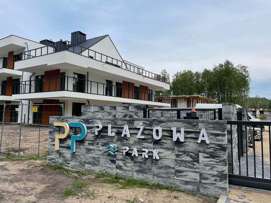 Nowoczesny budynek apartamentowy 'Plażowa Park' w trakcie budowy, widoczny przez kamienny mur z nazwą osiedla. Ujęcie z zewnątrz, w tle las.