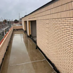 Wznoszenie ścian z cegły klinkierowej, widoczne czarne zabezpieczenie przeciwwilgociowe u podstawy muru. Na górze budynku widoczne rusztowania i dźwig.