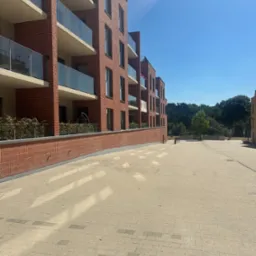 Nowoczesny budynek z czerwonej cegły z balkonami i szklanymi balustradami w słoneczny dzień. Widoczna elewacja i chodnik z kostki.