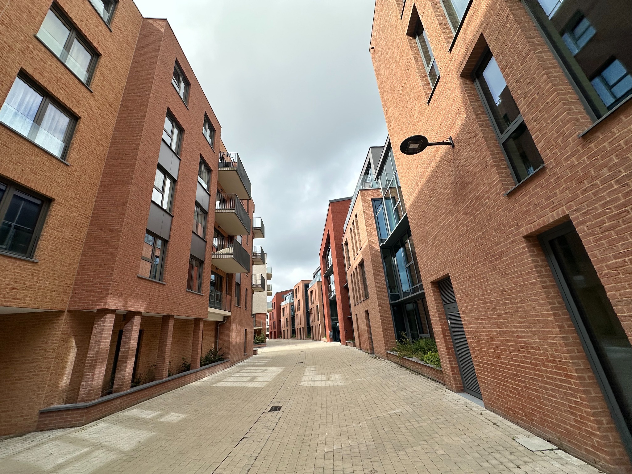 Ulica z budynkami z czerwonej cegły w nowoczesnym stylu. Widok z poziomu chodnika, perspektywa zbieżna, pochmurne niebo. Geometryczne detale architektoniczne.