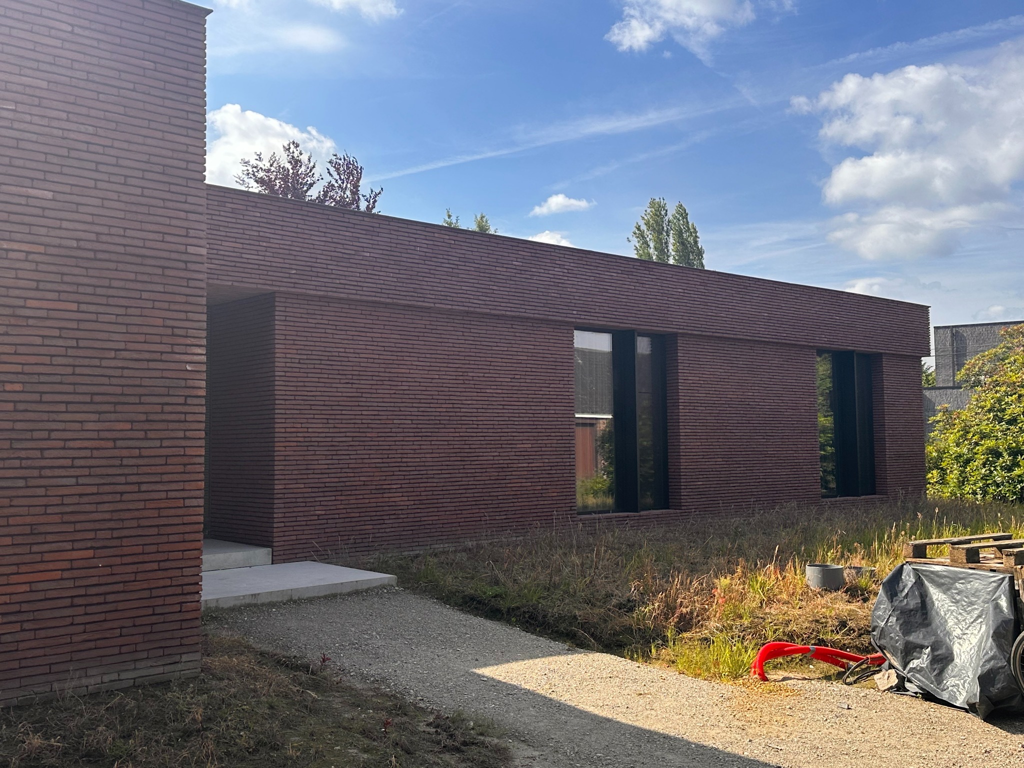 Nowoczesny dom z ciemnej cegły klinkierowej, z minimalistycznymi oknami i betonową ścieżką. Widoczne materiały budowlane i otoczenie w trakcie wykończenia.