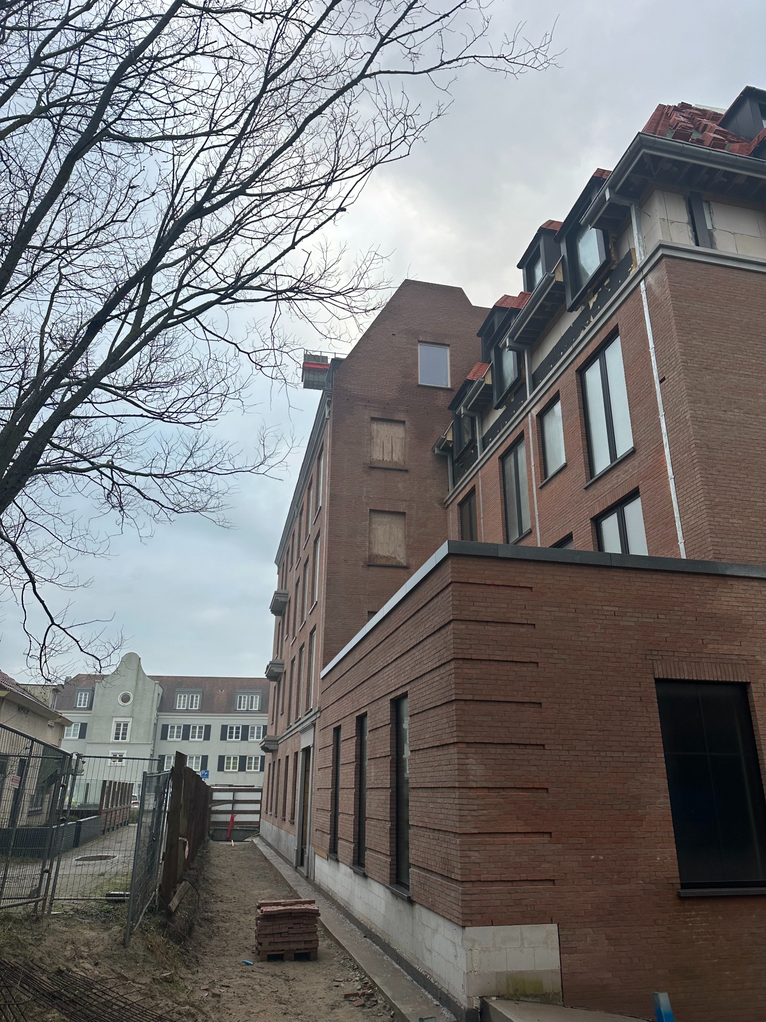 Budynek z czerwonej cegły w trakcie budowy, widoczne okna, dach z czerwonej dachówki, rusztowania i materiały budowlane. Na pierwszym planie drzewo bez liści.