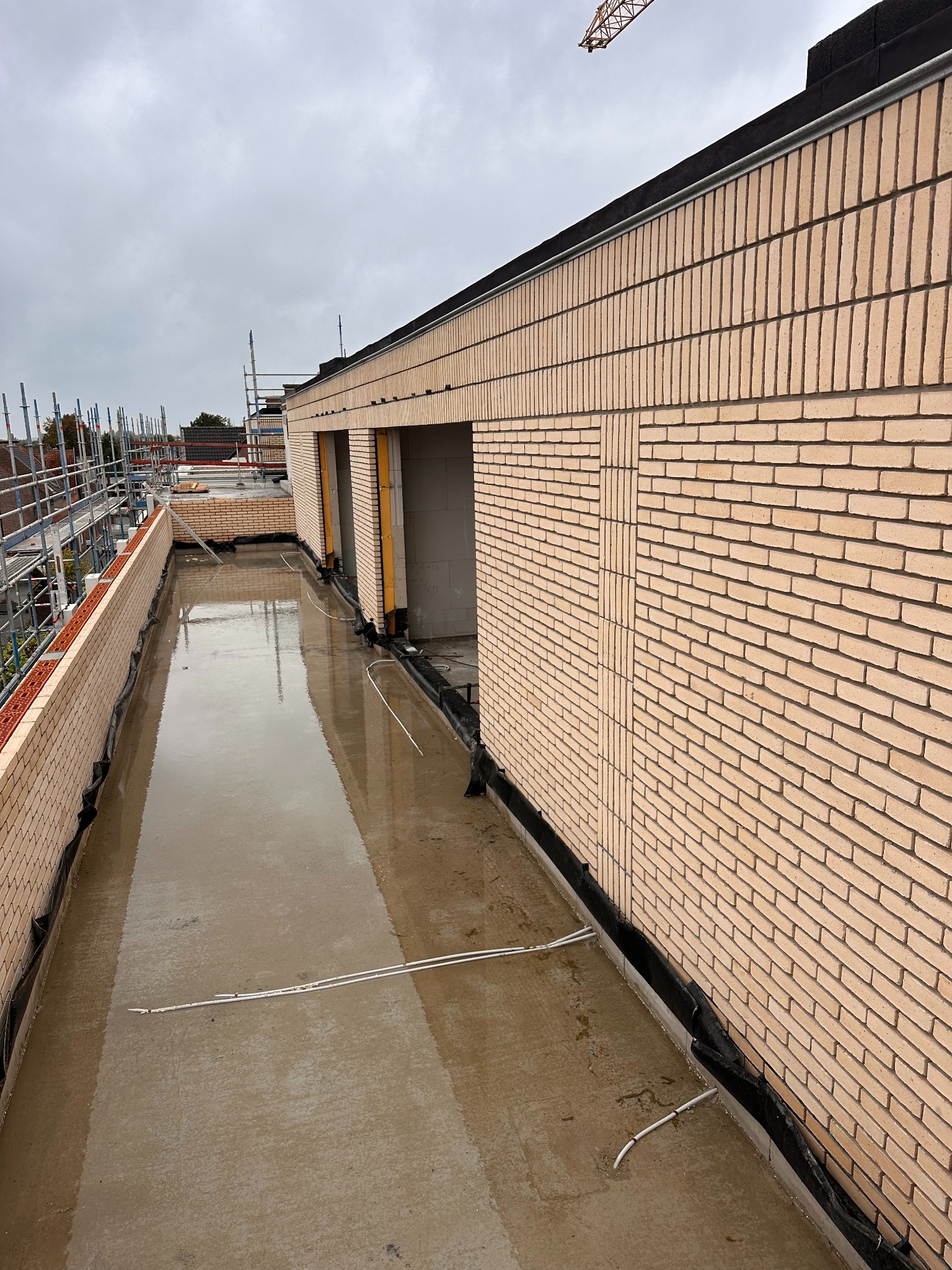 Wznoszenie ścian z cegły klinkierowej, widoczne czarne zabezpieczenie przeciwwilgociowe u podstawy muru. Na górze budynku widoczne rusztowania i dźwig.