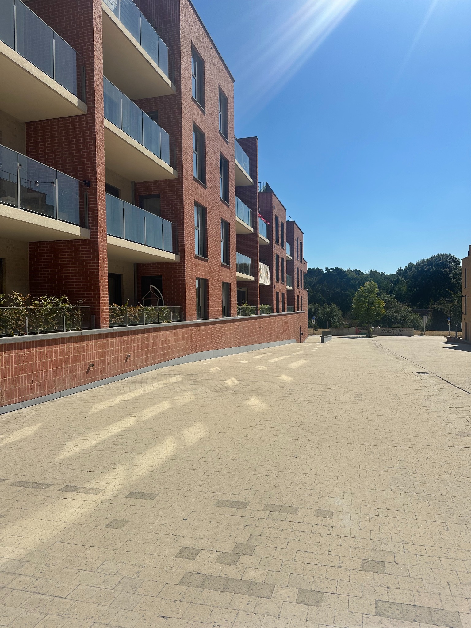 Nowoczesny budynek z czerwonej cegły z balkonami i szklanymi balustradami w słoneczny dzień. Widoczna elewacja i chodnik z kostki.