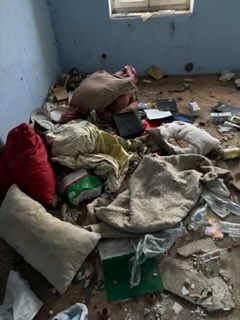 Zaniedbane wnętrze pokoju z chaotycznym nagromadzeniem przedmiotów, w tym poduszek, worków i śmieci, na tle niebieskich ścian i okna.