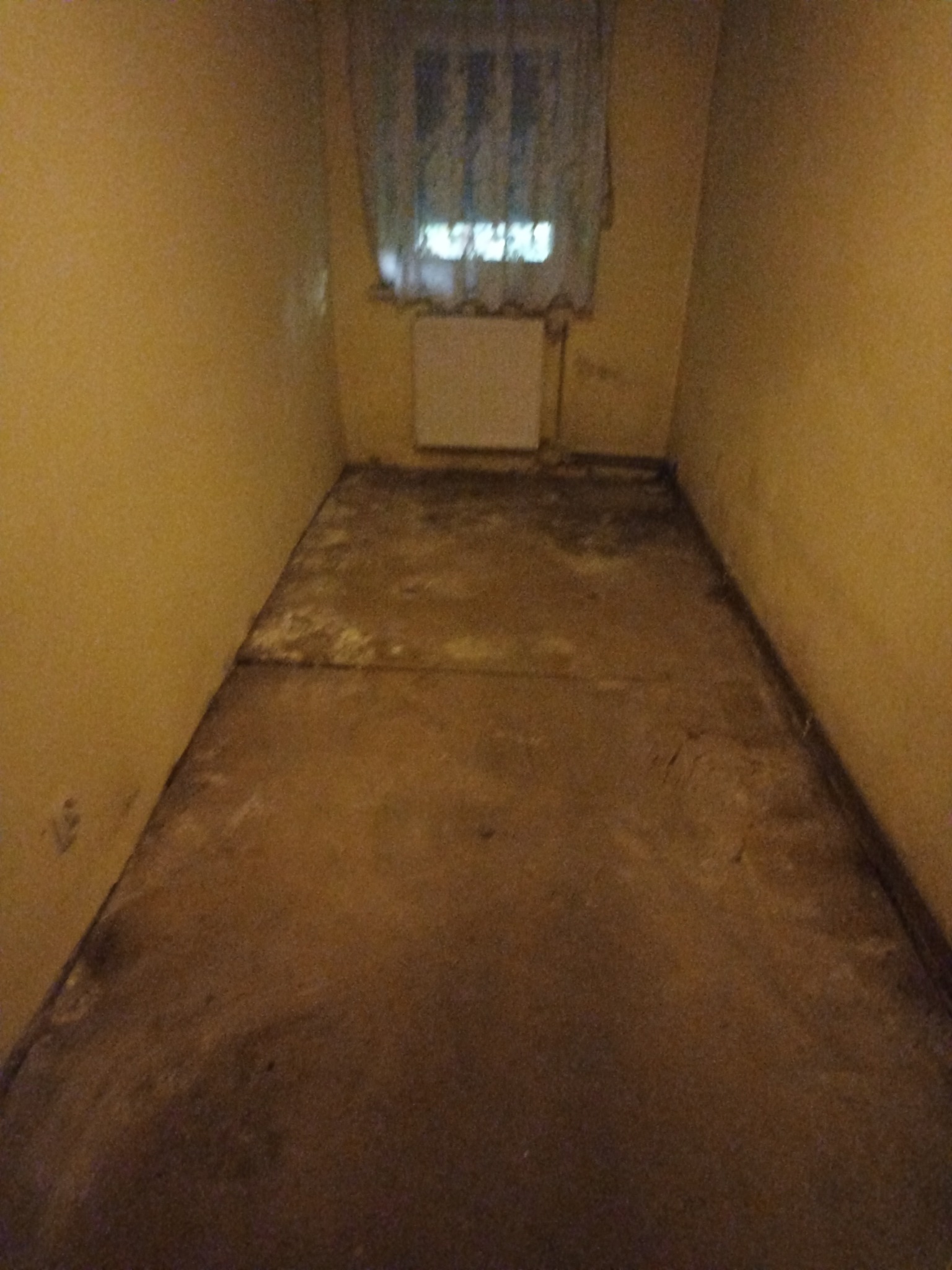 Pokój w mieszkaniu po opróżnieniu, widoczne ślady po meblach na podłodze i ścianach. Okno z firanką w tle. Żółte ściany i betonowa podłoga.