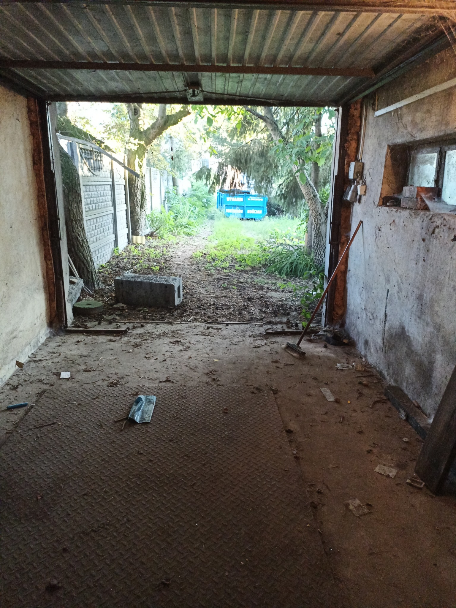 Zaniedbany garaż z podłogą z blachy ryflowanej, śmieciami i widokiem na zarośnięte podwórze. Stara miotła oparta o ścianę, betonowe elementy w otworze bramy.