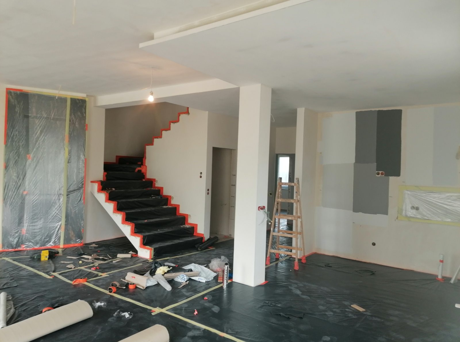 Wnętrze w trakcie prac wykończeniowych: zabezpieczone schody, folia ochronna na podłodze, częściowo pomalowana ściana z widocznymi puszkami elektrycznymi.