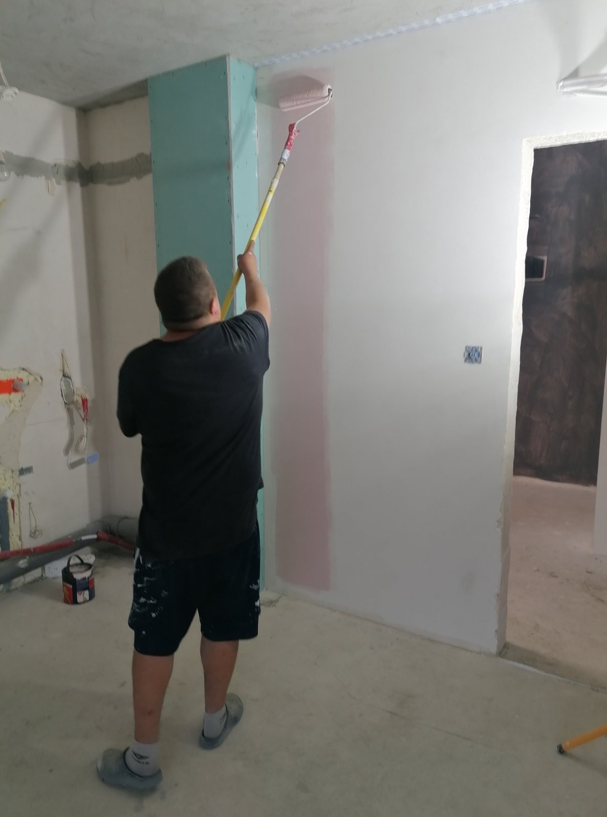 Malowanie ścian wałkiem w pomieszczeniu z zabudową z płyt. Widoczny fragment ściany pomalowany na różowo i narzędzia malarskie. Prace wykończeniowe.