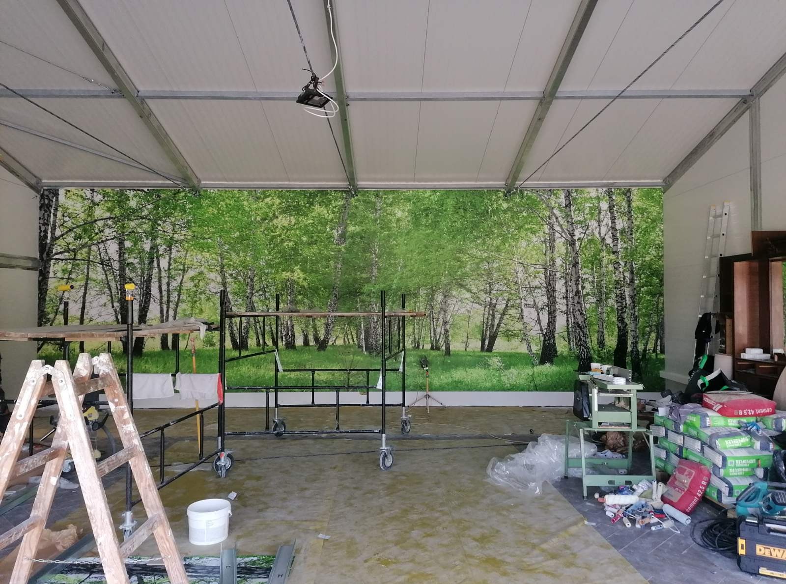 Montaż okładziny ściennej z motywem leśnym wewnątrz hali. Widoczne rusztowania, drabina i narzędzia sugerują przygotowanie do dalszych prac wykończeniowych.