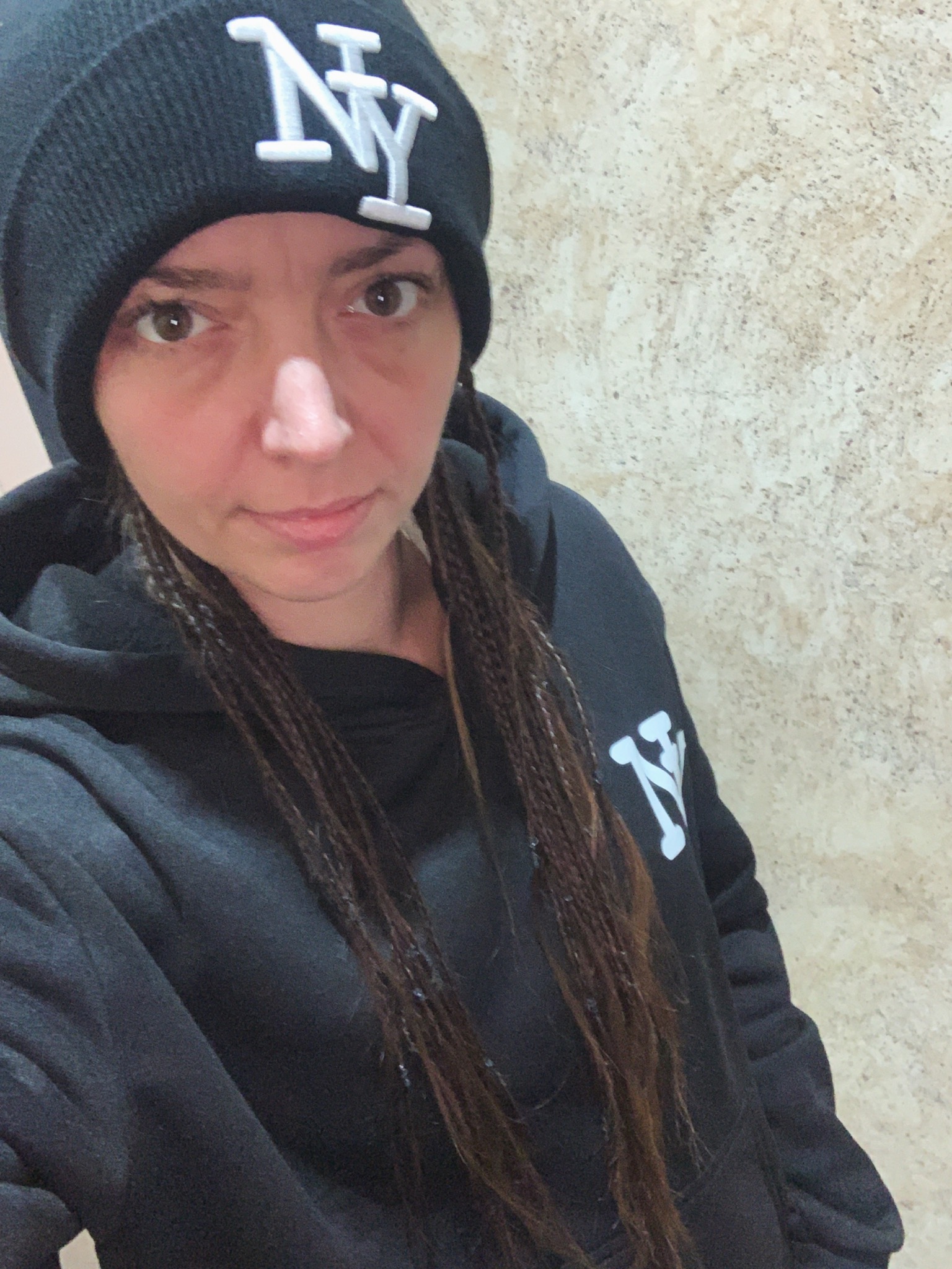 Portret kobiety w czarnej bluzie z kapturem i czapce z logo NY, z warkoczykami, na tle ściany o fakturze imitującej kamień. Ujęcie z bliska, oświetlenie naturalne.